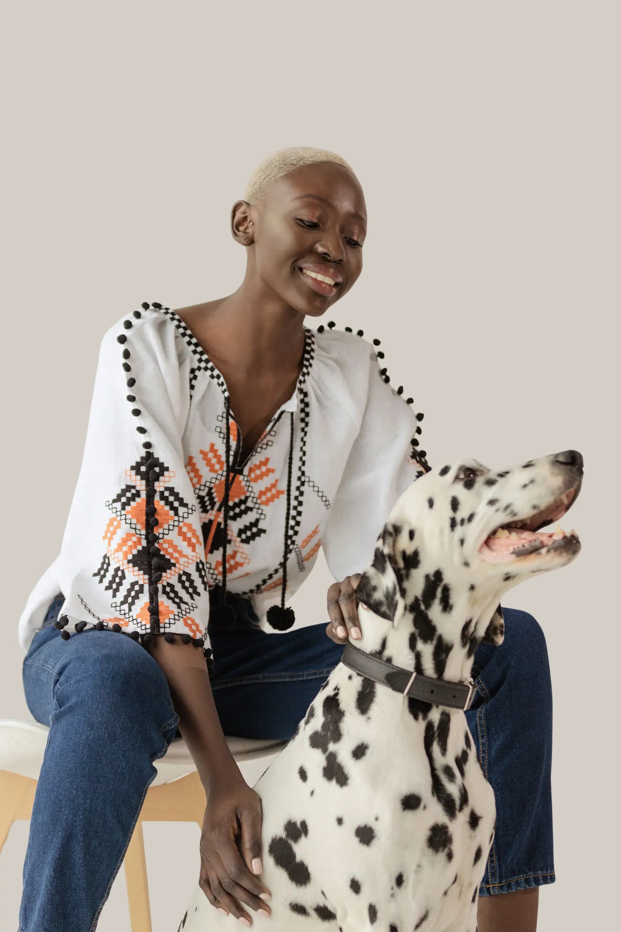 A woman with short blonde hair sits on a stool wearing a white embroidered blouse with black and orange geometric patterns, blue jeans, and a black pendant necklace. She is smiling and petting a Dalmatian dog wearing a black collar, against a solid light beige background.