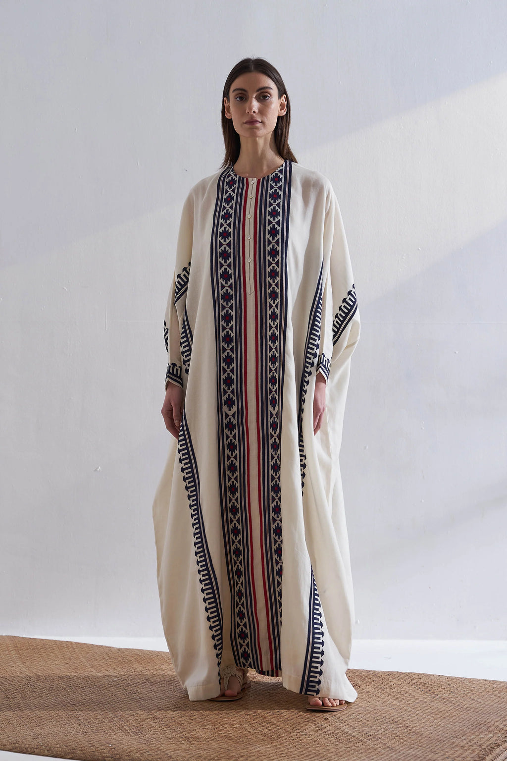Woman wearing a long, cream-colored dress with black and red patterns on a plain background