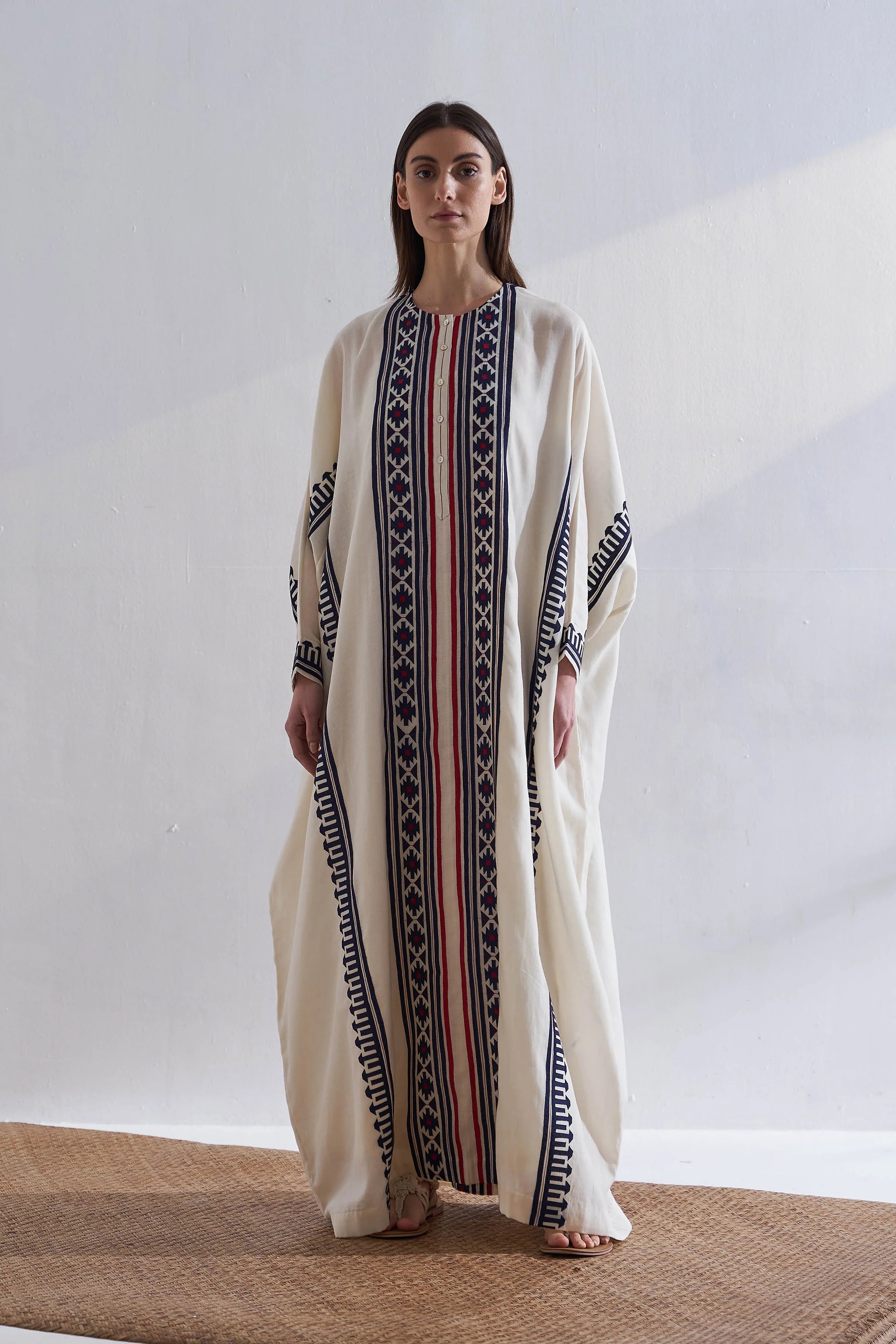 Woman wearing a long, cream-colored dress with black and red patterns on a plain background