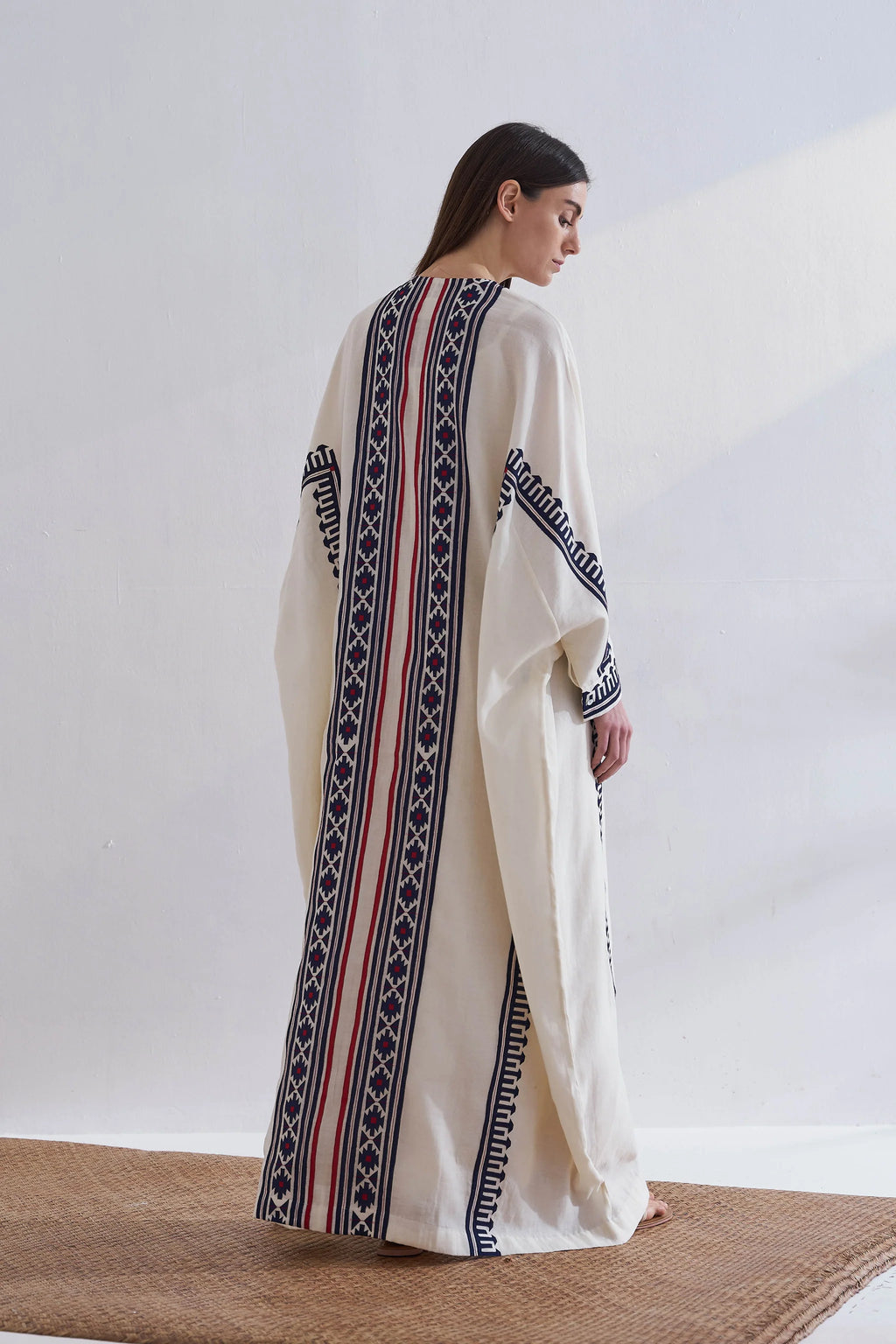 A model in a cream-colored long kaftan featuring vertical navy blue geometric appliqué panels and red accent stripes.