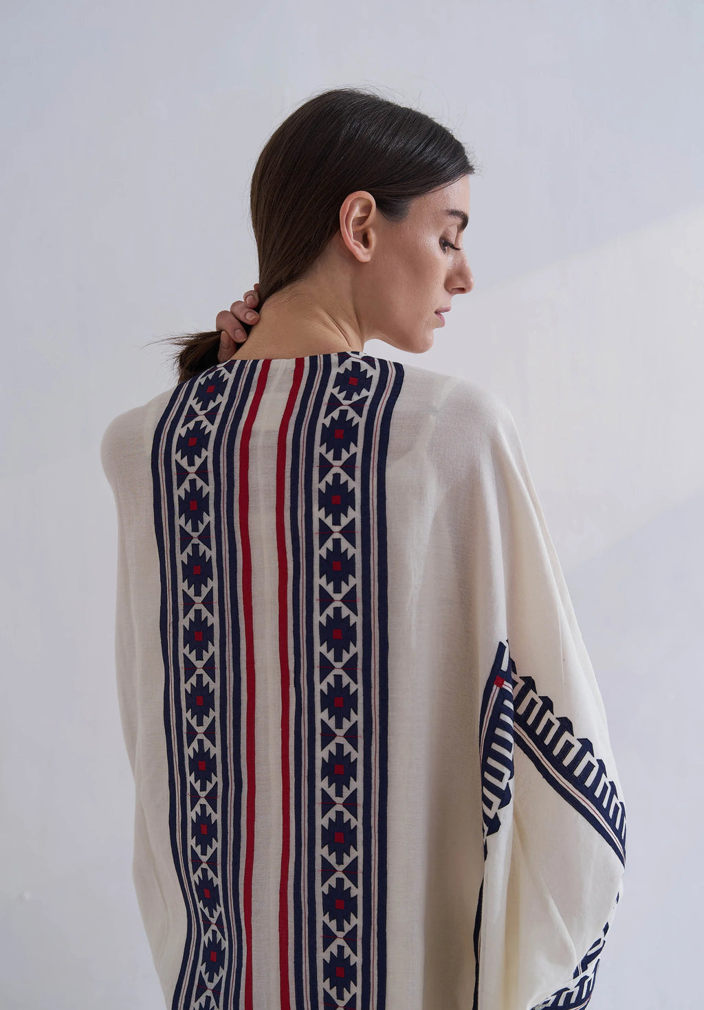 A model in a cream-colored long kaftan featuring vertical navy blue geometric appliqué panels and red accent stripes.