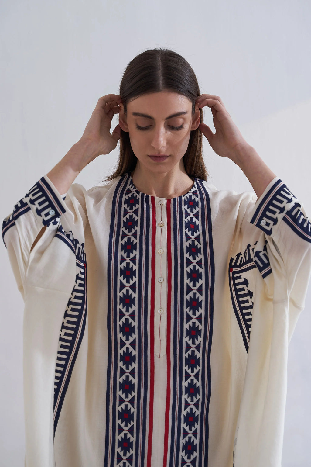 A model in a cream-colored long kaftan featuring vertical navy blue geometric appliqué panels and red accent stripes.