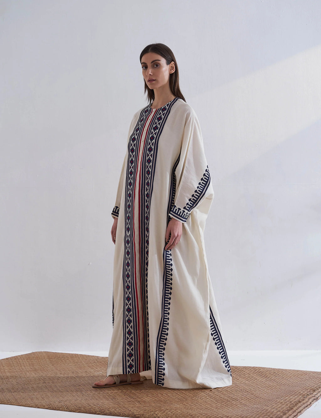 A model in a cream-colored long kaftan featuring vertical navy blue geometric appliqué panels and red accent stripes.