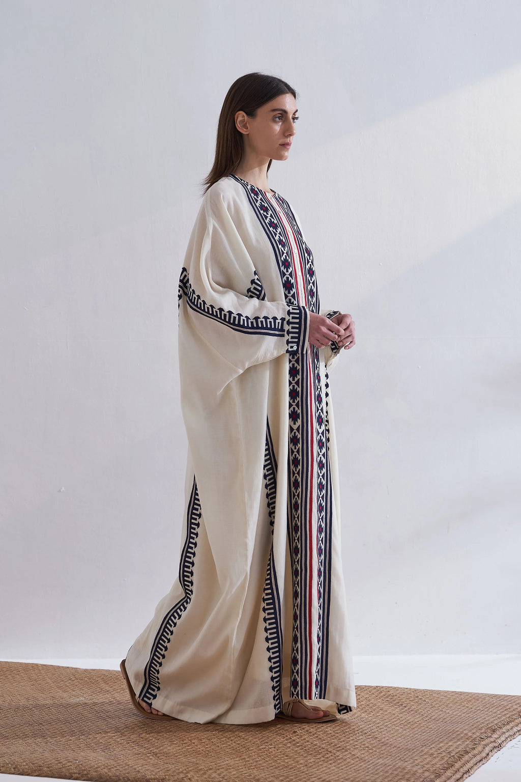 A model in a cream-colored long kaftan featuring vertical navy blue geometric appliqué panels and red accent stripes.