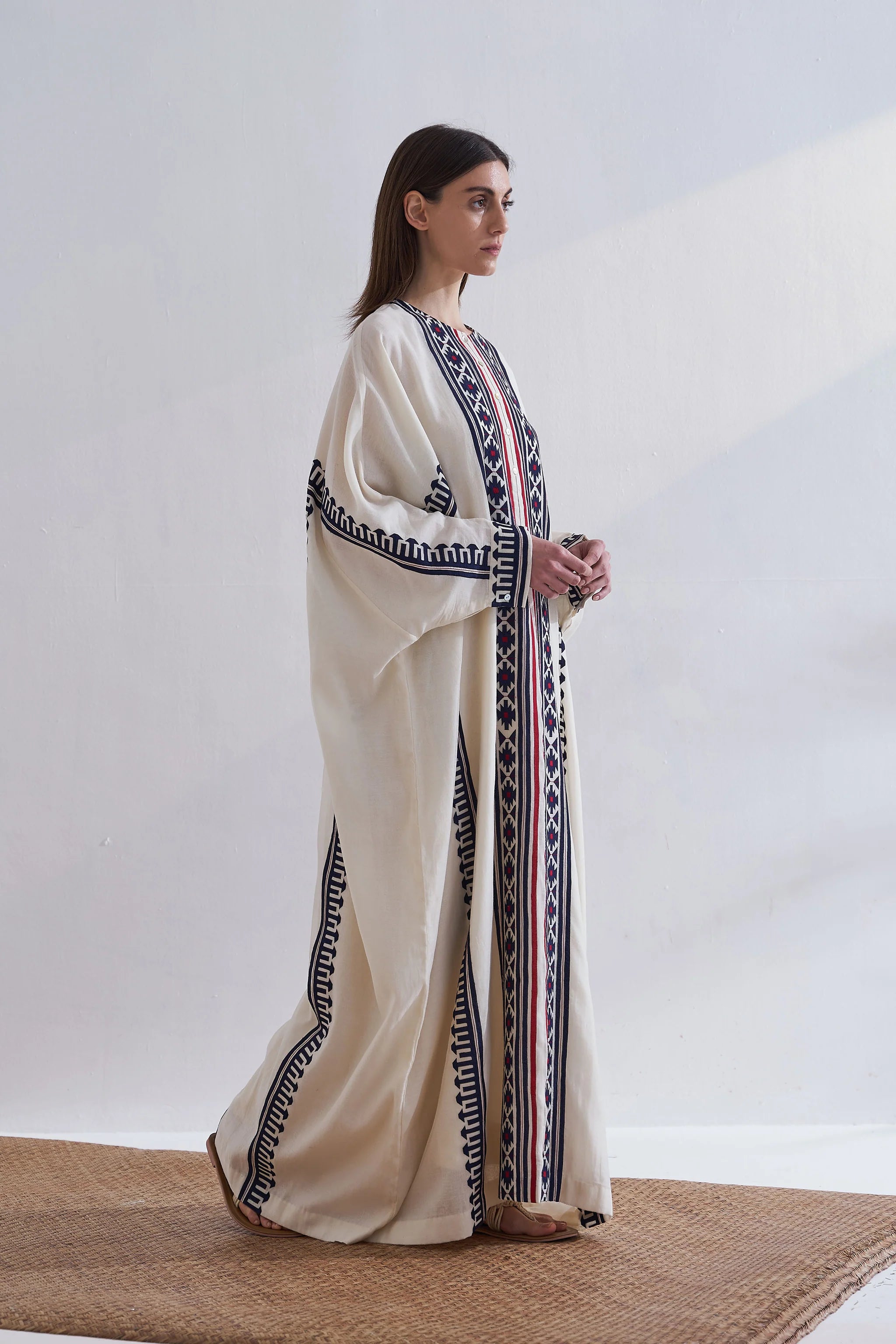 A model in a cream-colored long kaftan featuring vertical navy blue geometric appliqué panels and red accent stripes.