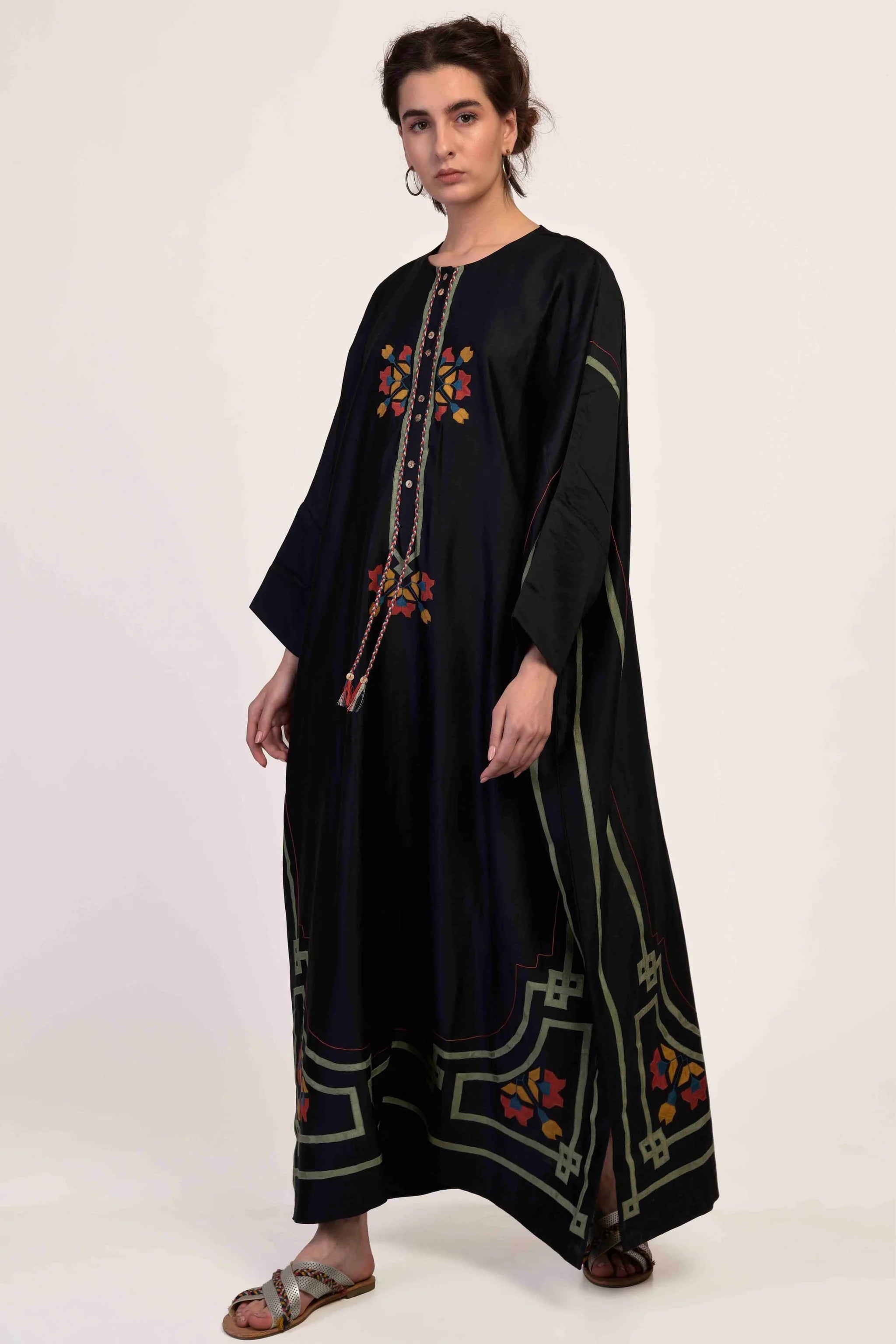 A model in a floor-length black kaftan with intricate geometric hand-cut appliqué in green, red, and yellow tones.