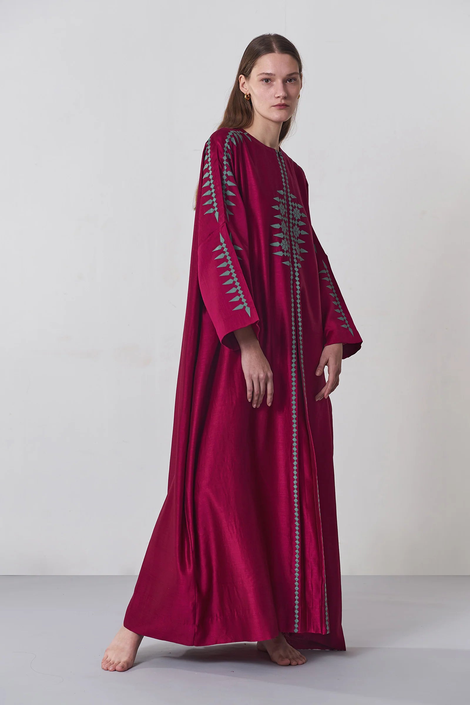 A model wearing a deep maroon long kaftan dress featuring sage green tribal-inspired hand-cut appliqué down the center and sleeves.