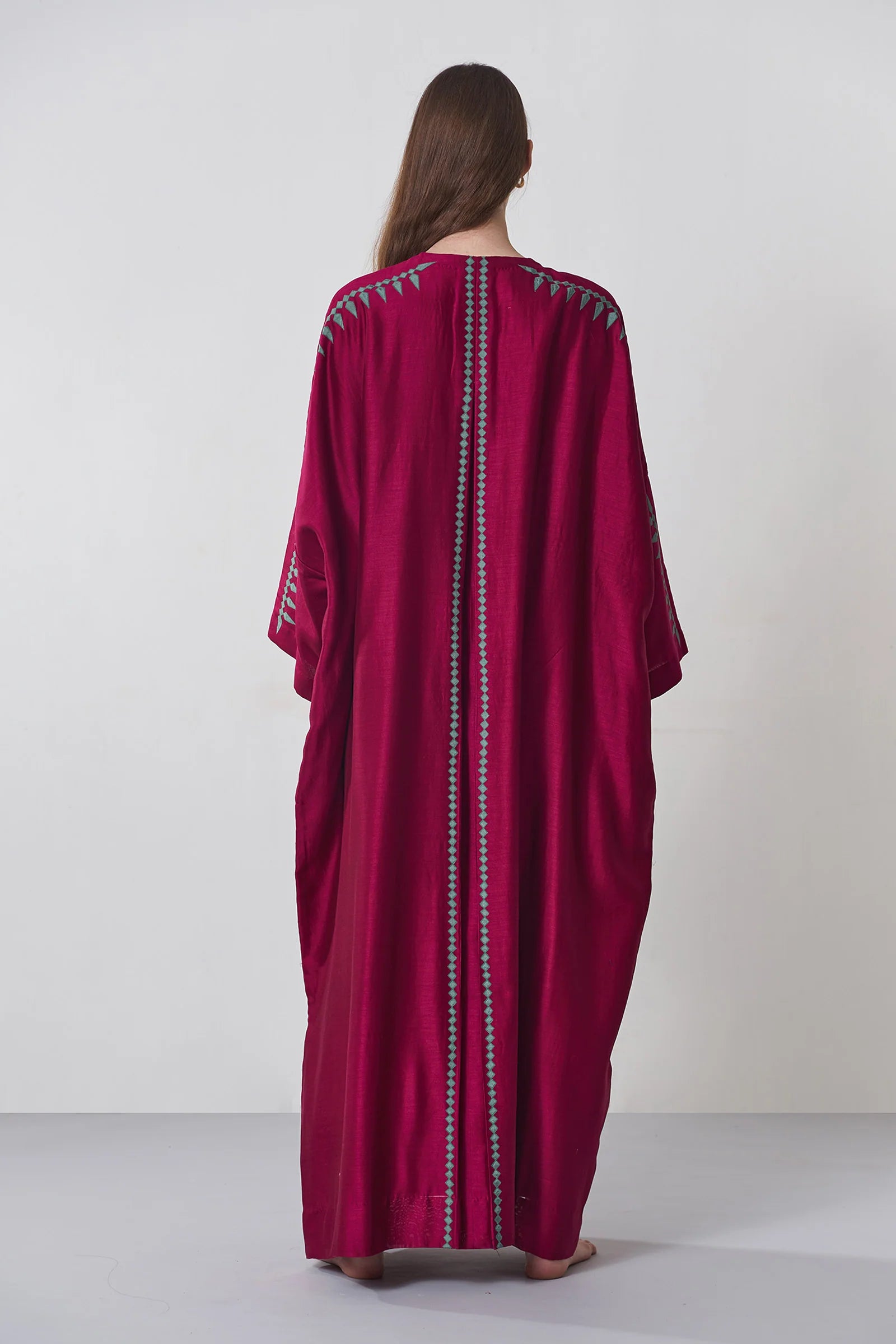 A model wearing a deep maroon long kaftan dress featuring sage green tribal-inspired hand-cut appliqué down the center and sleeves.