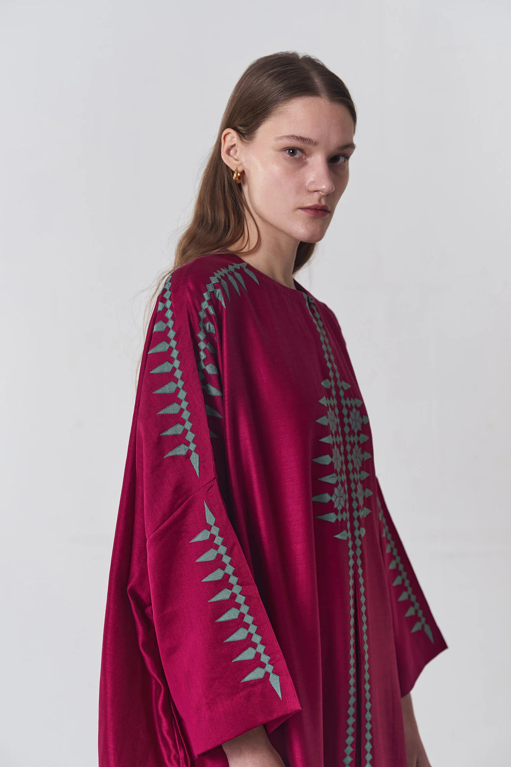 A model wearing a deep maroon long kaftan dress featuring sage green tribal-inspired hand-cut appliqué down the center and sleeves.