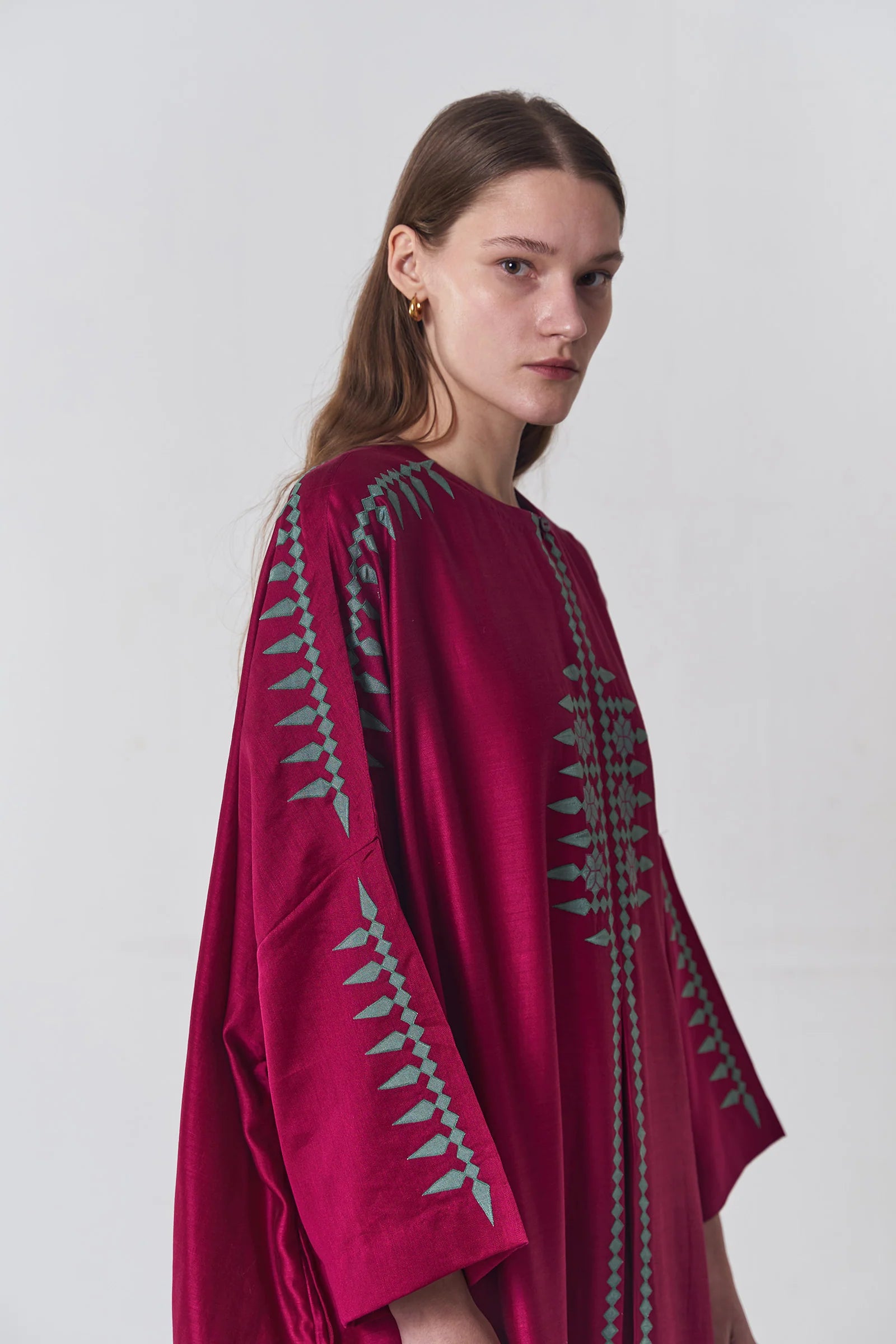A model wearing a deep maroon long kaftan dress featuring sage green tribal-inspired hand-cut appliqué down the center and sleeves.