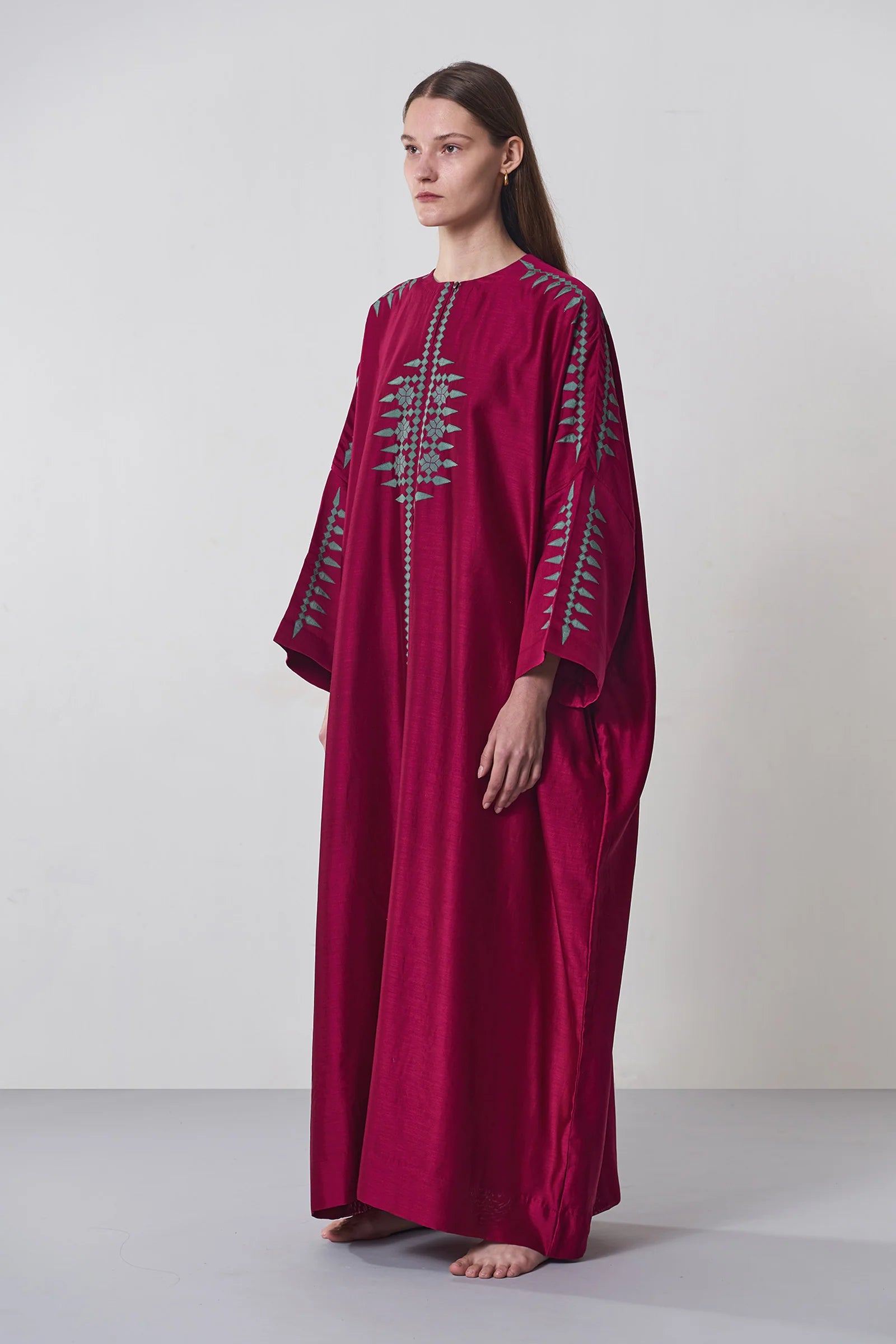 A model wearing a deep maroon long kaftan dress featuring sage green tribal-inspired hand-cut appliqué down the center and sleeves.