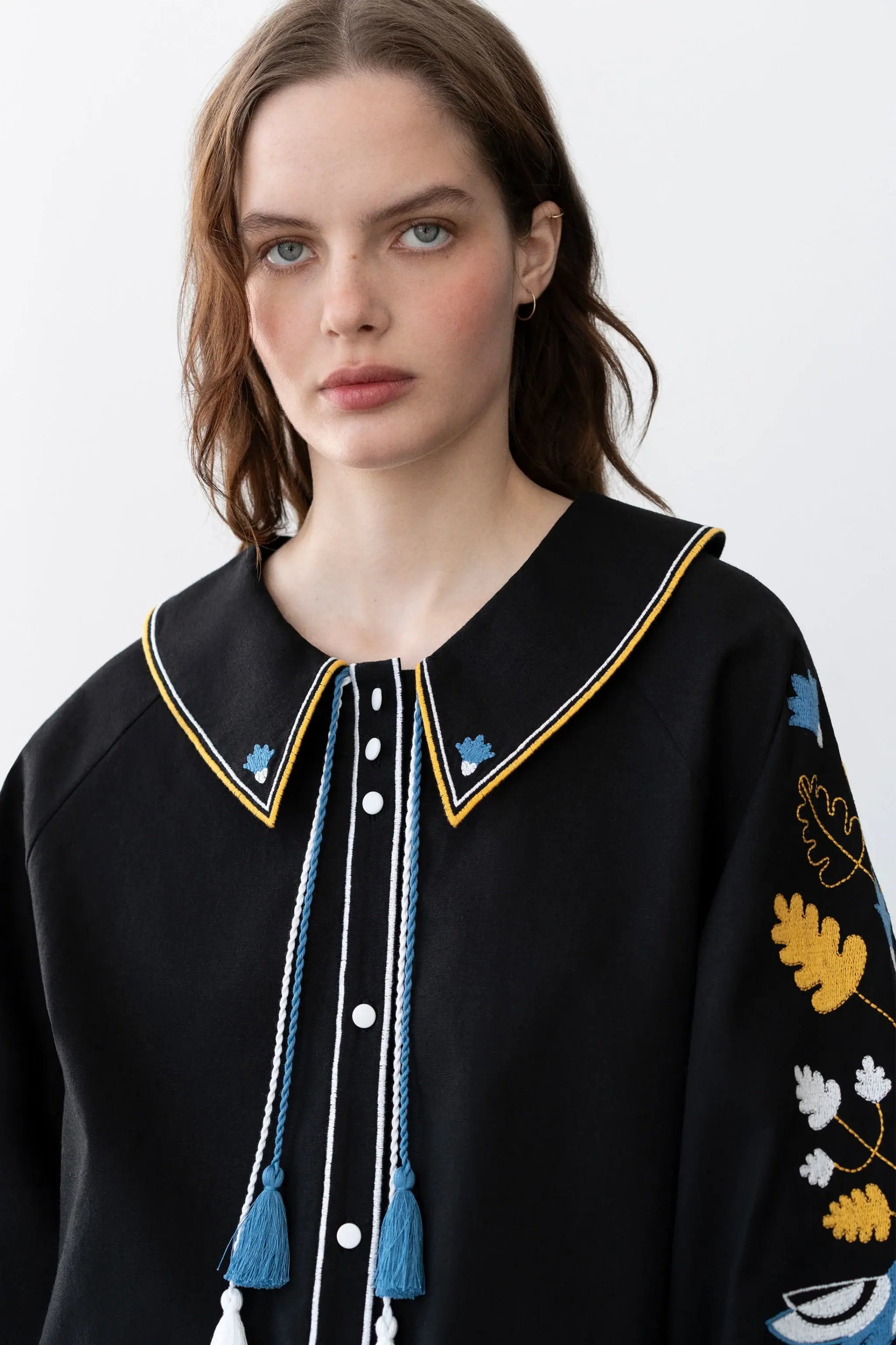 A close-up of a woman wearing a black embroidered top with a pointed collar, blue and yellow trim, tassel details, and floral embroidery on the sleeve.