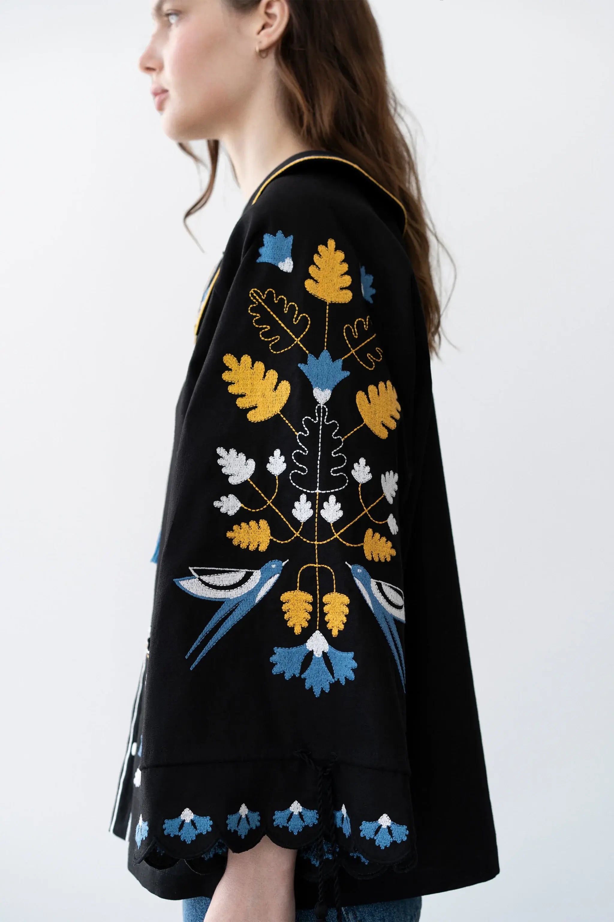A woman shown in side profile wearing a black embroidered jacket with blue, yellow, and white floral and bird motifs, photographed against a plain light background.