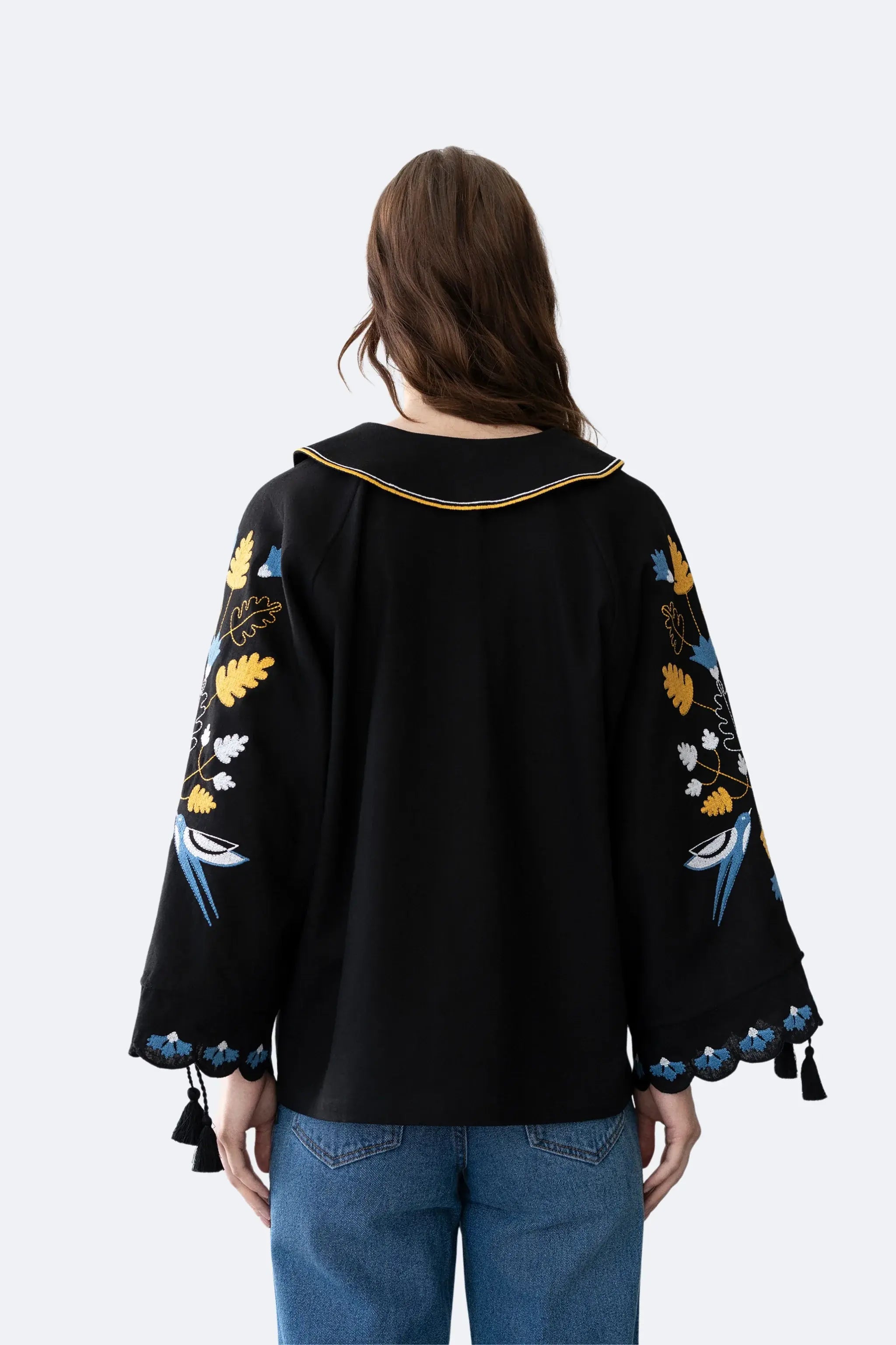 A woman viewed from the back wearing a black long-sleeve blouse with colorful embroidered floral and bird motifs on the sleeves, blue jeans, and a white collar with a thin gold trim, against a light gray studio background.