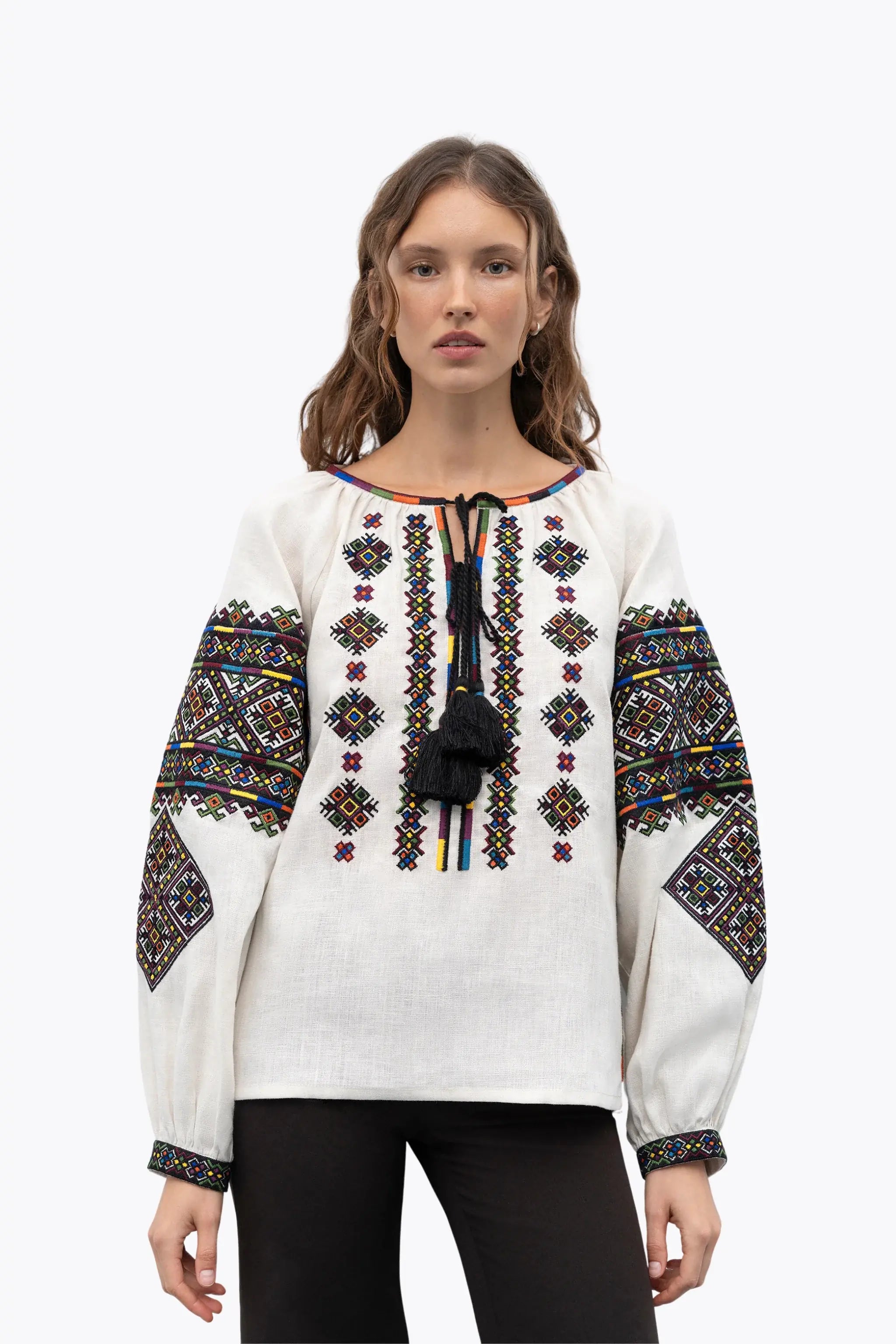 A woman modeling a white embroidered blouse with colorful geometric patterns on the chest and sleeves, black tassels at the neckline, and black pants, shot against a plain white background.