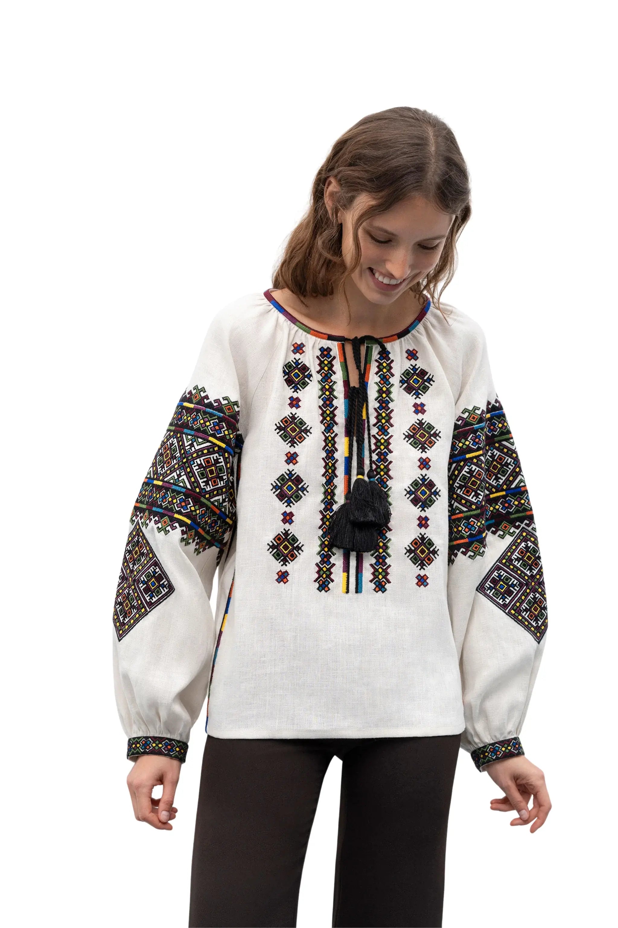 A woman wearing a white long-sleeve blouse with colorful geometric embroidery on the chest and sleeves, paired with black pants, photographed against a plain white background.