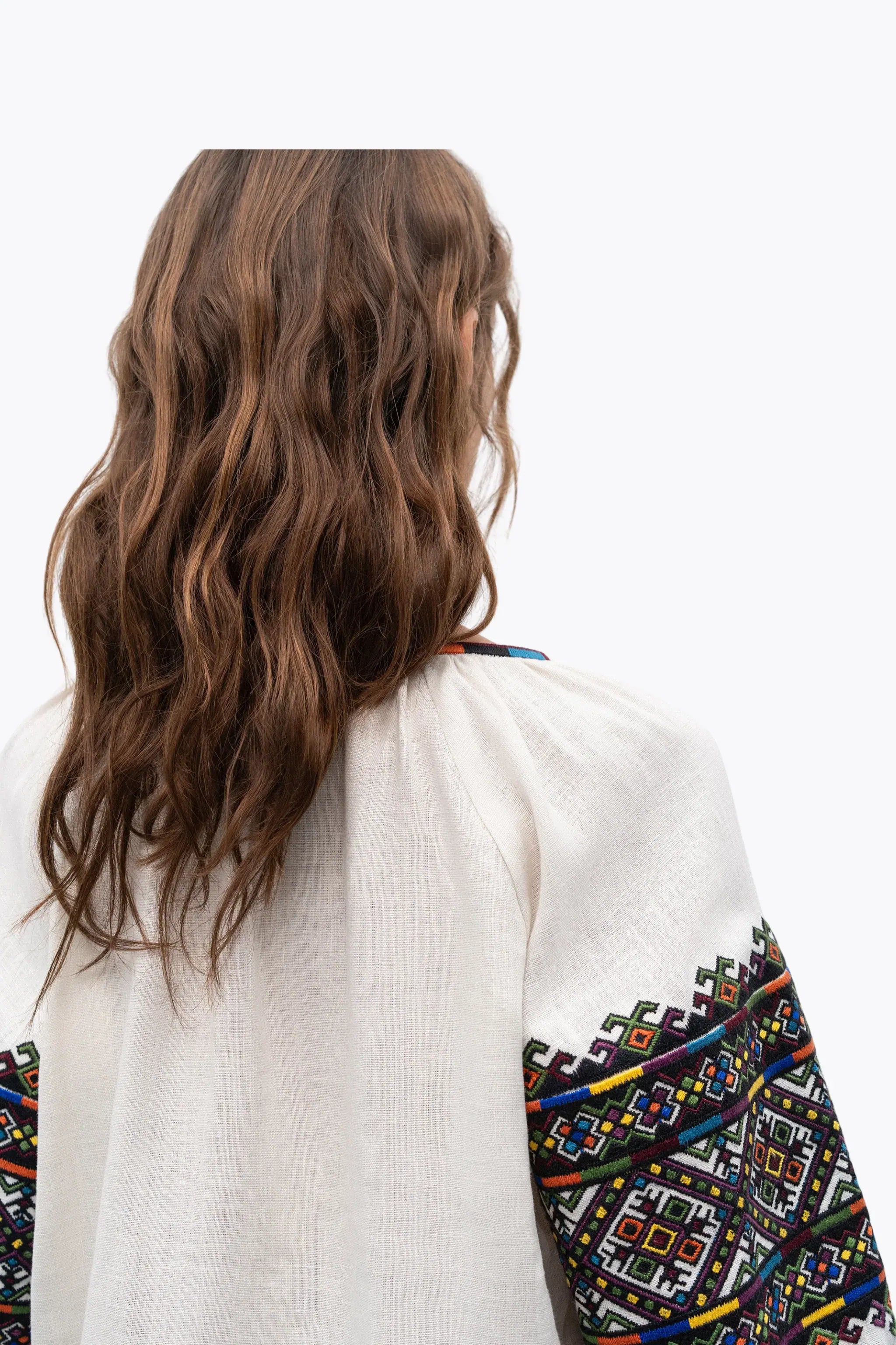 A woman viewed from behind wearing a white linen blouse with colorful geometric embroidered patterns on the sleeves, set against a plain white background.