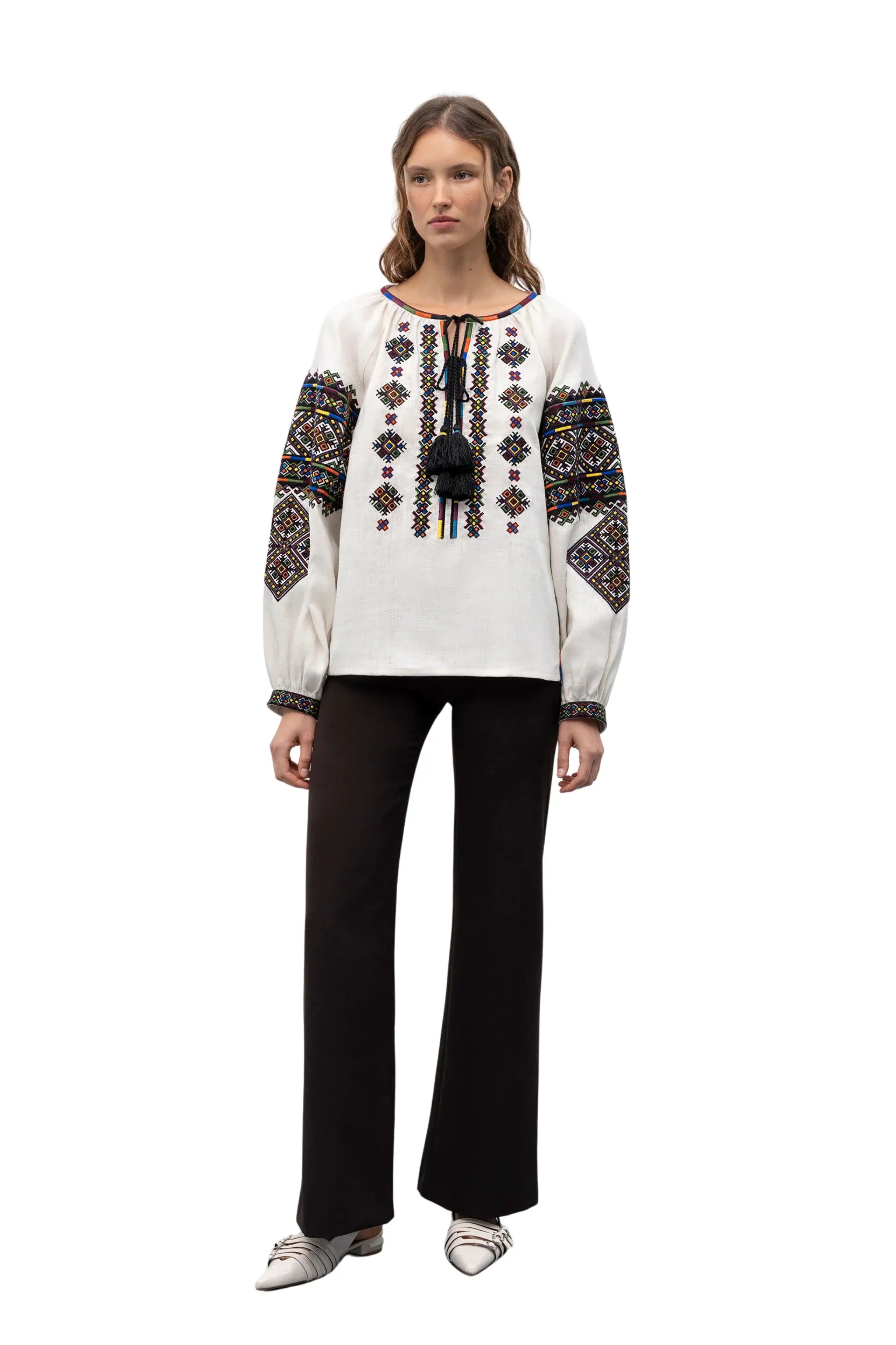 A model wearing a white long-sleeve embroidered blouse with colorful geometric patterns on the sleeves and chest, paired with black wide-leg pants and white pointed flats, set against a plain white background.