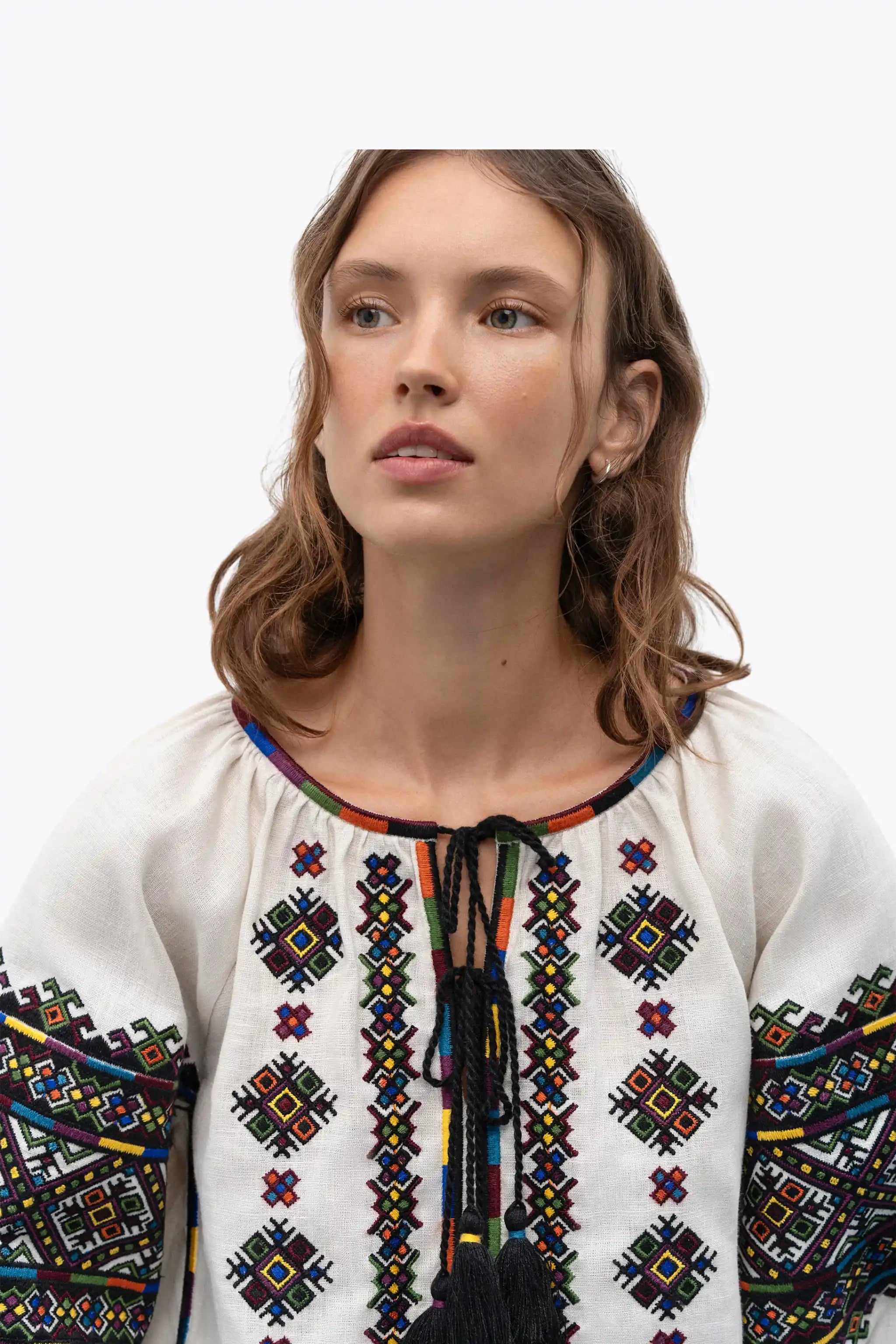 A woman wearing a white embroidered blouse with colorful geometric patterns and black tassel ties at the neckline, shown against a plain light gray background.
