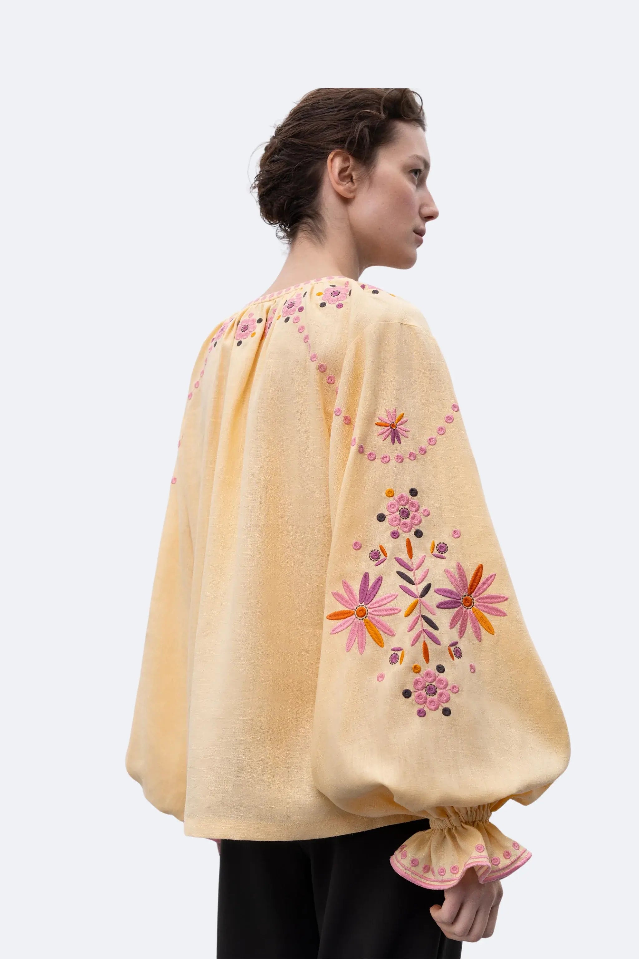 A woman shown from the back wearing a pale yellow embroidered cape-style top with voluminous sleeves and colorful floral embroidery, paired with black pants, against a light gray studio background.