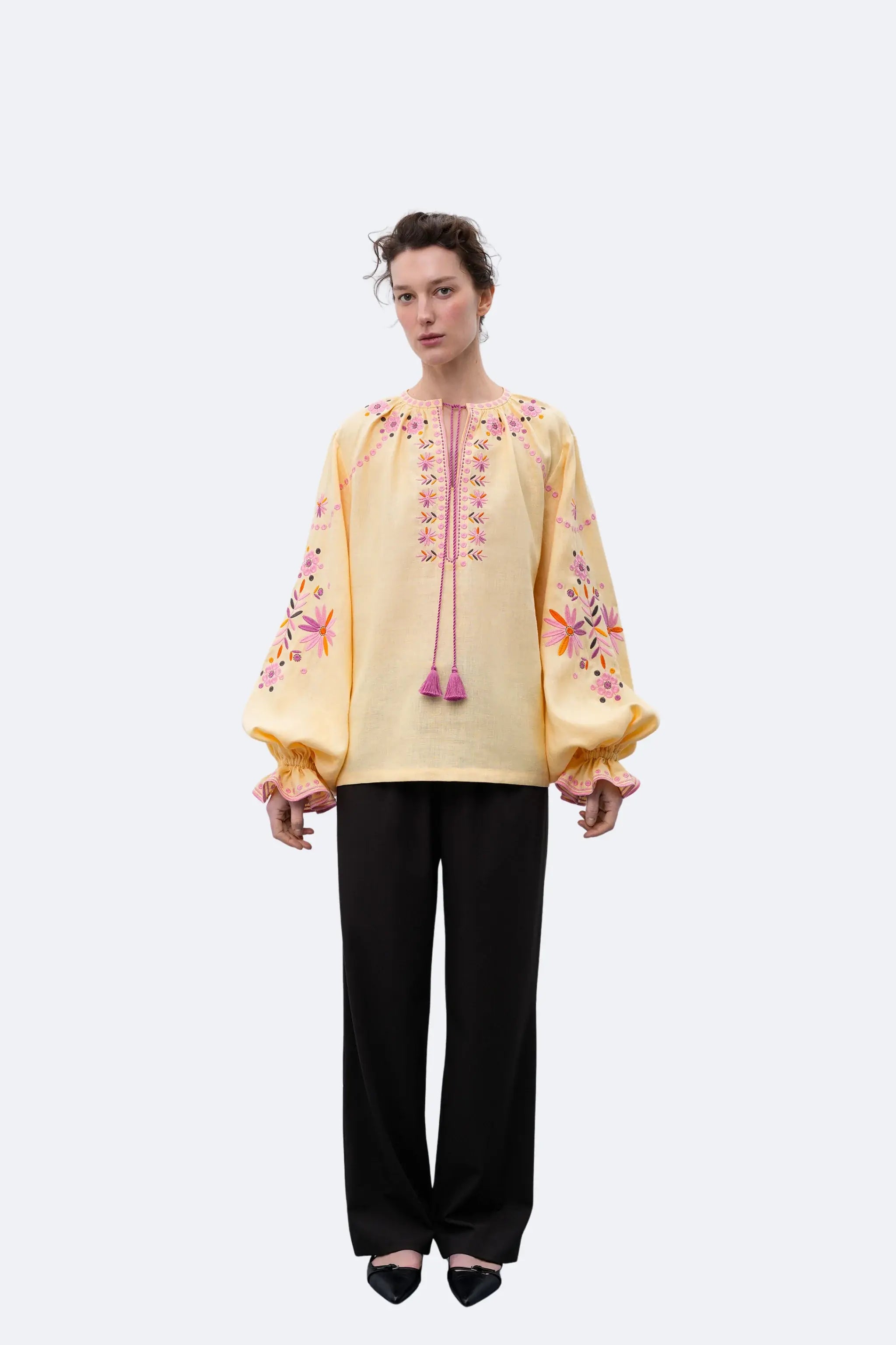 A model stands in profile wearing a pale yellow embroidered blouse with pink and orange floral detailing and tassel ties, paired with black wide-leg trousers and black pointed flats, against a light gray studio background.