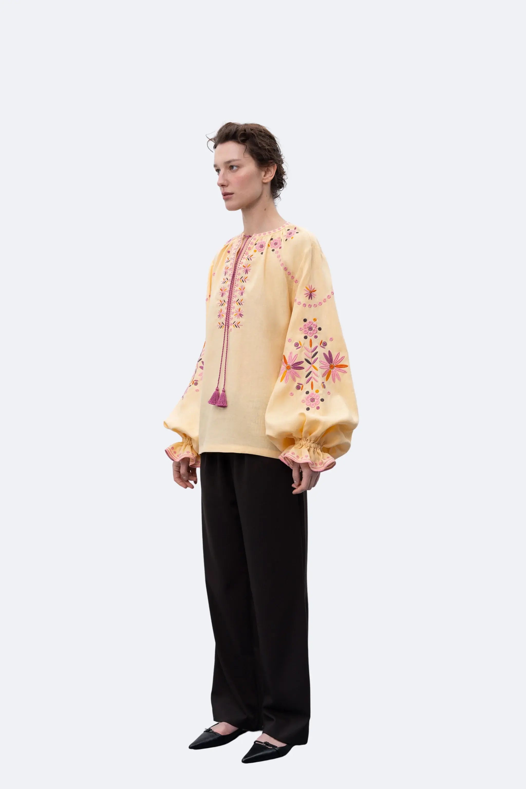 A model stands in profile wearing a pale yellow embroidered blouse with pink and orange floral detailing and tassel ties, paired with black wide-leg trousers and black pointed flats, against a light gray studio background.