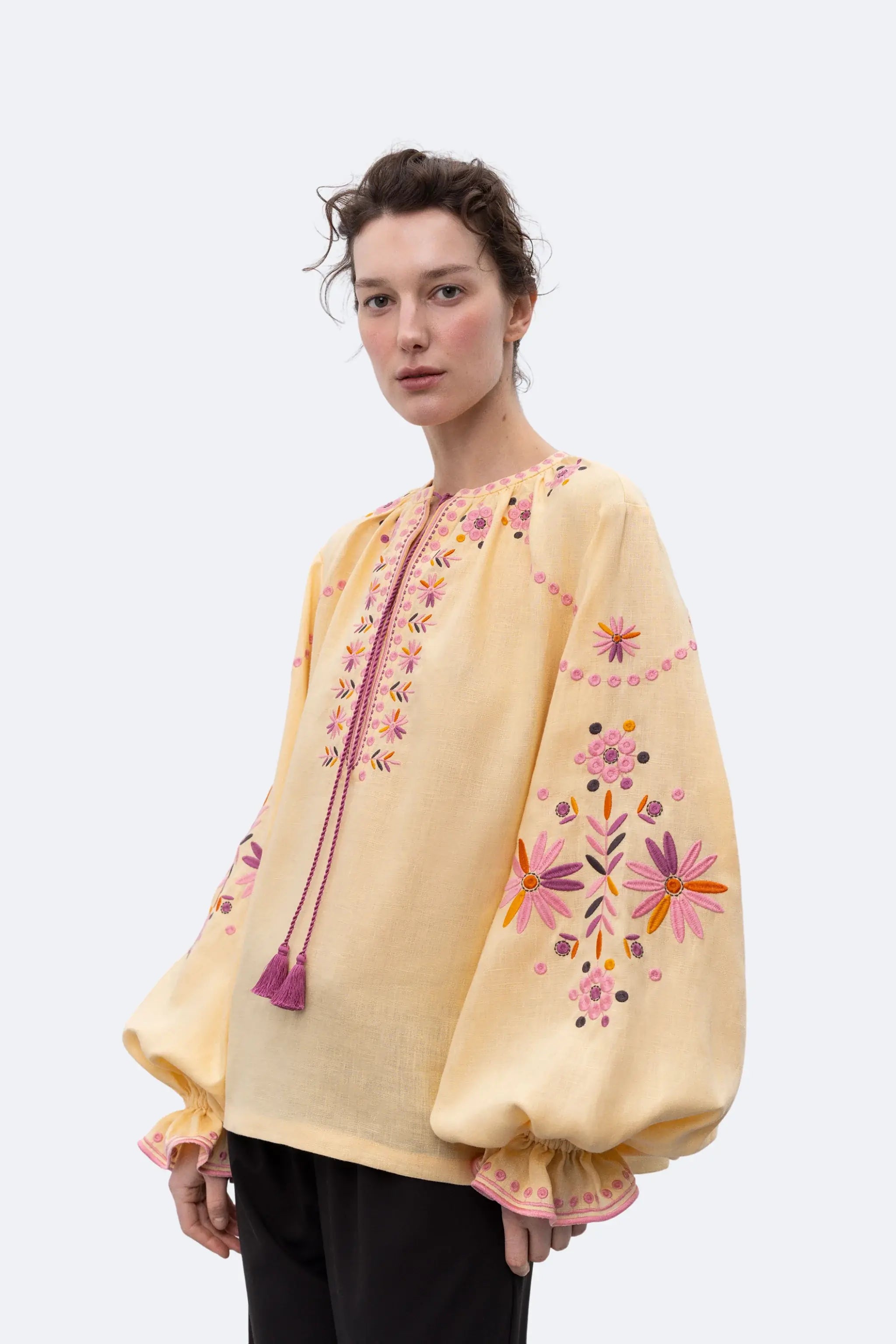 A model stands in profile wearing a pale yellow embroidered blouse with pink and orange floral detailing and tassel ties, paired with black wide-leg trousers and black pointed flats, against a light gray studio background.