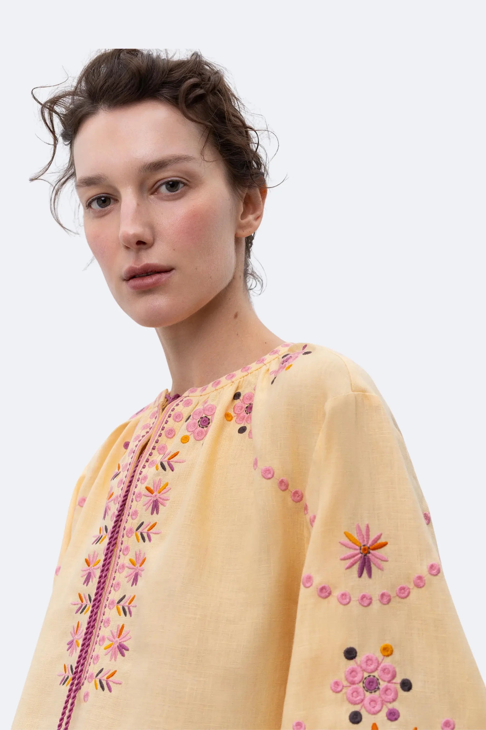 A model stands in profile wearing a pale yellow embroidered blouse with pink and orange floral detailing and tassel ties, paired with black wide-leg trousers and black pointed flats, against a light gray studio background.