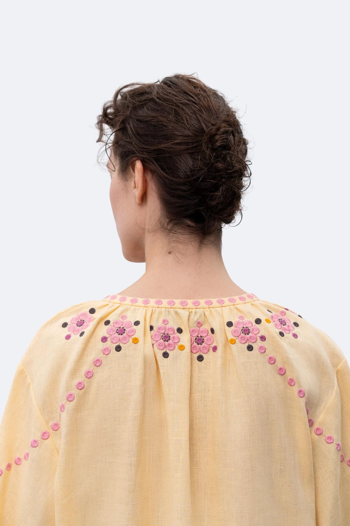 A model stands in profile wearing a pale yellow embroidered blouse with pink and orange floral detailing and tassel ties, paired with black wide-leg trousers and black pointed flats, against a light gray studio background.