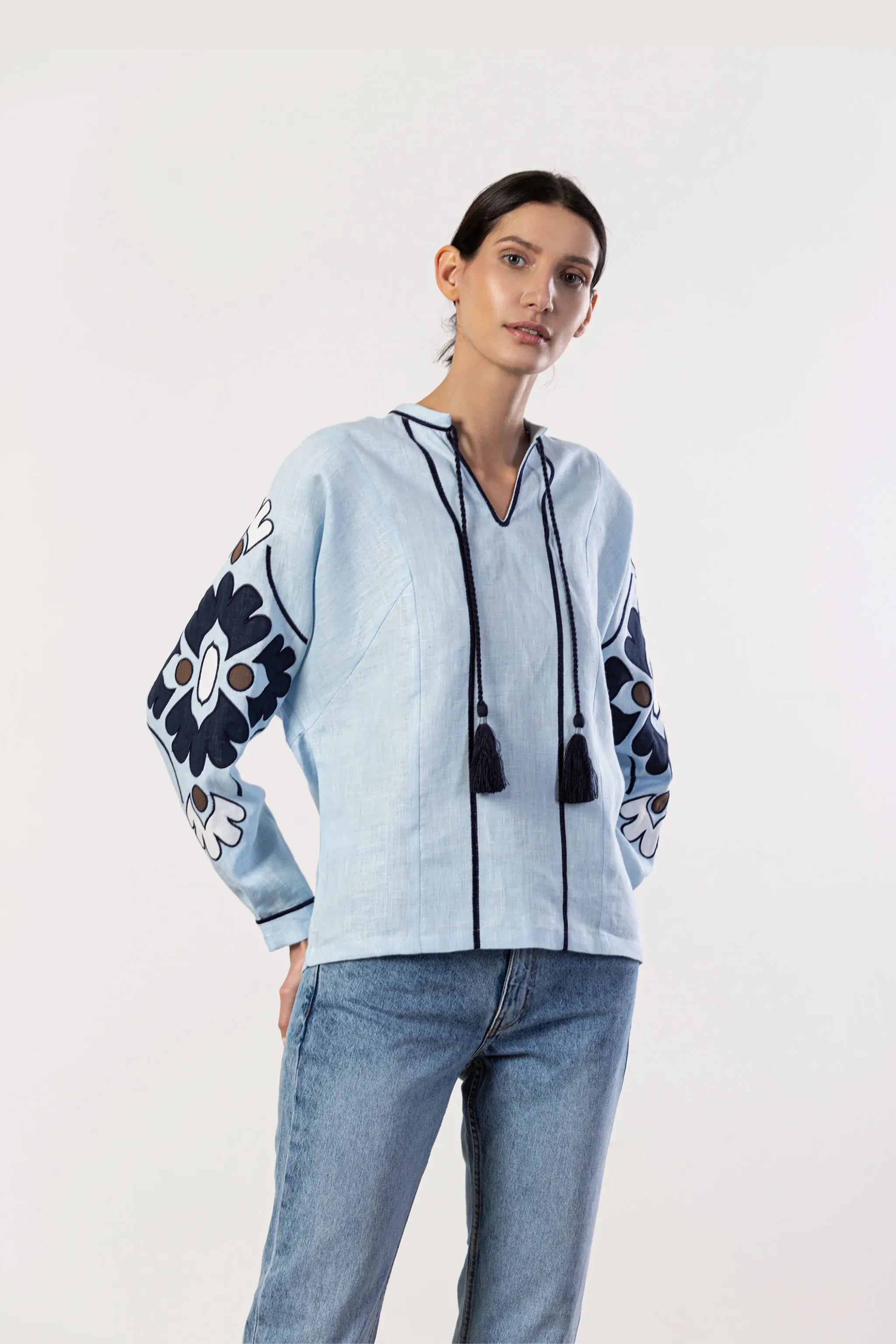 A woman modeling a light blue embroidered long-sleeve top with dark navy floral patterns on the sleeves and black tassel drawstrings, paired with blue jeans against a plain white studio background.