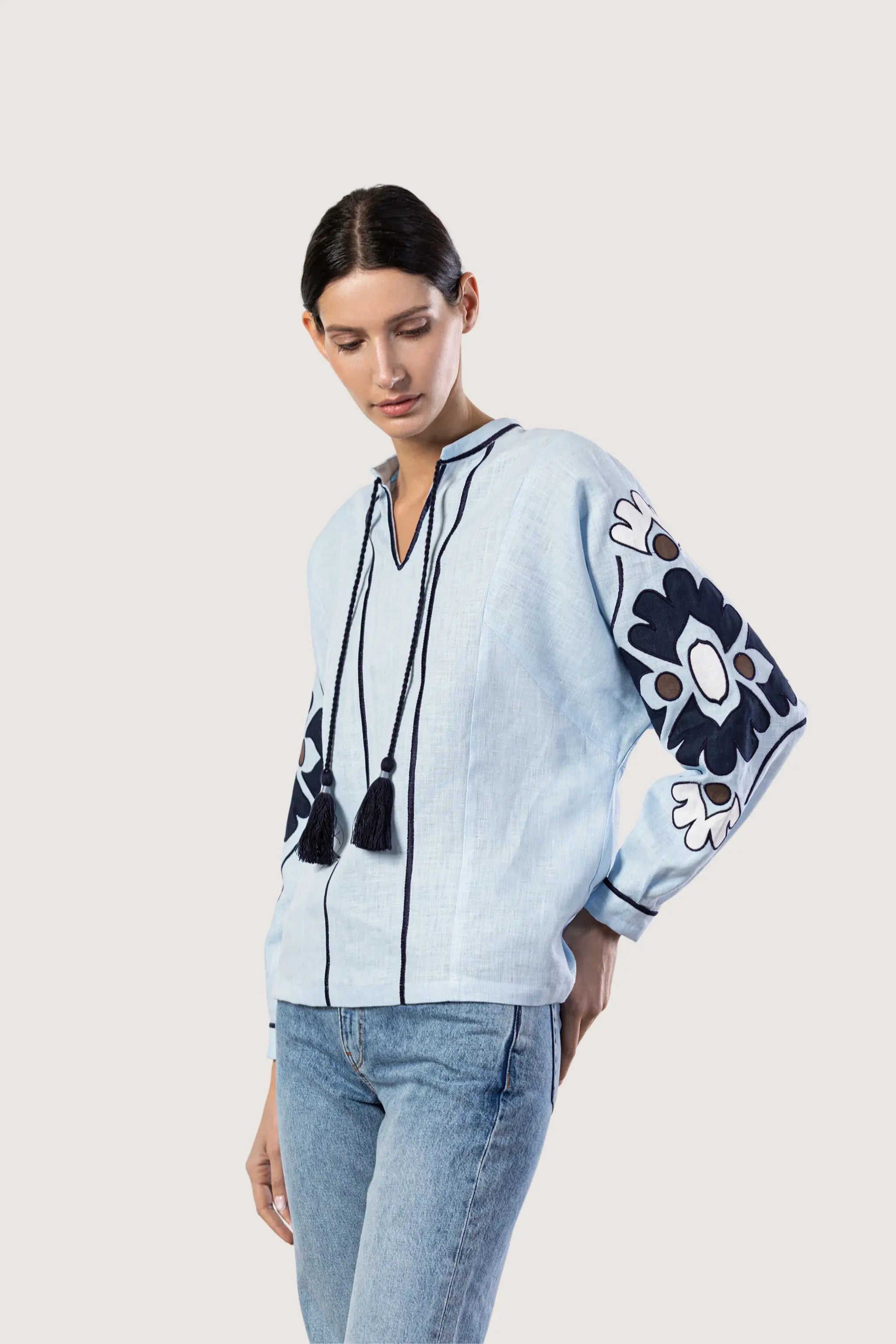 A woman modeling a light blue embroidered blouse with dark navy floral sleeve patterns and tassel necklaces, paired with blue jeans, against a solid light gray background.