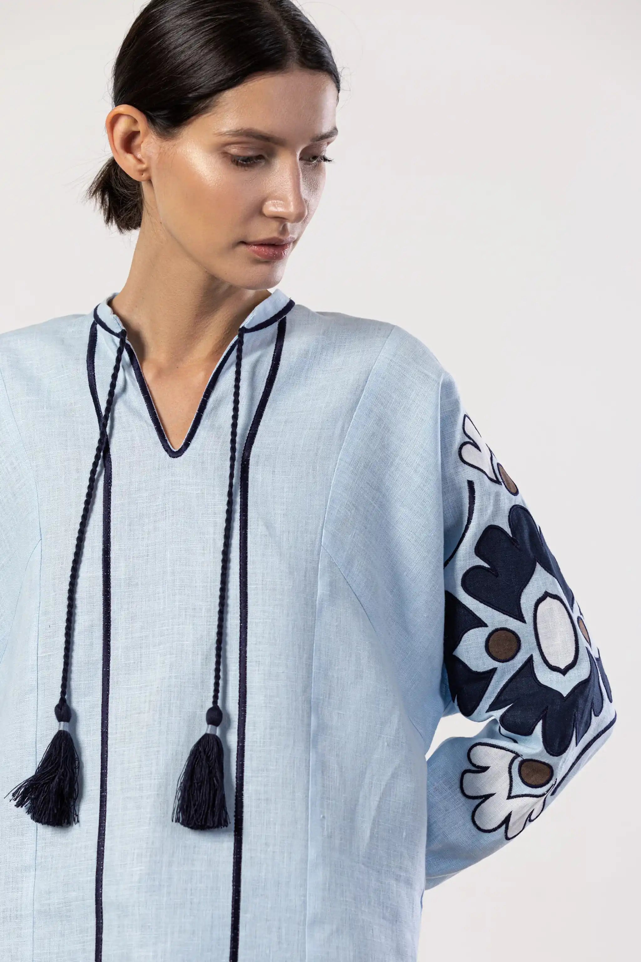 A woman wearing a light blue linen tunic top with dark navy blue trim, tassels, and floral embroidery on the sleeve, set against a plain light gray background.