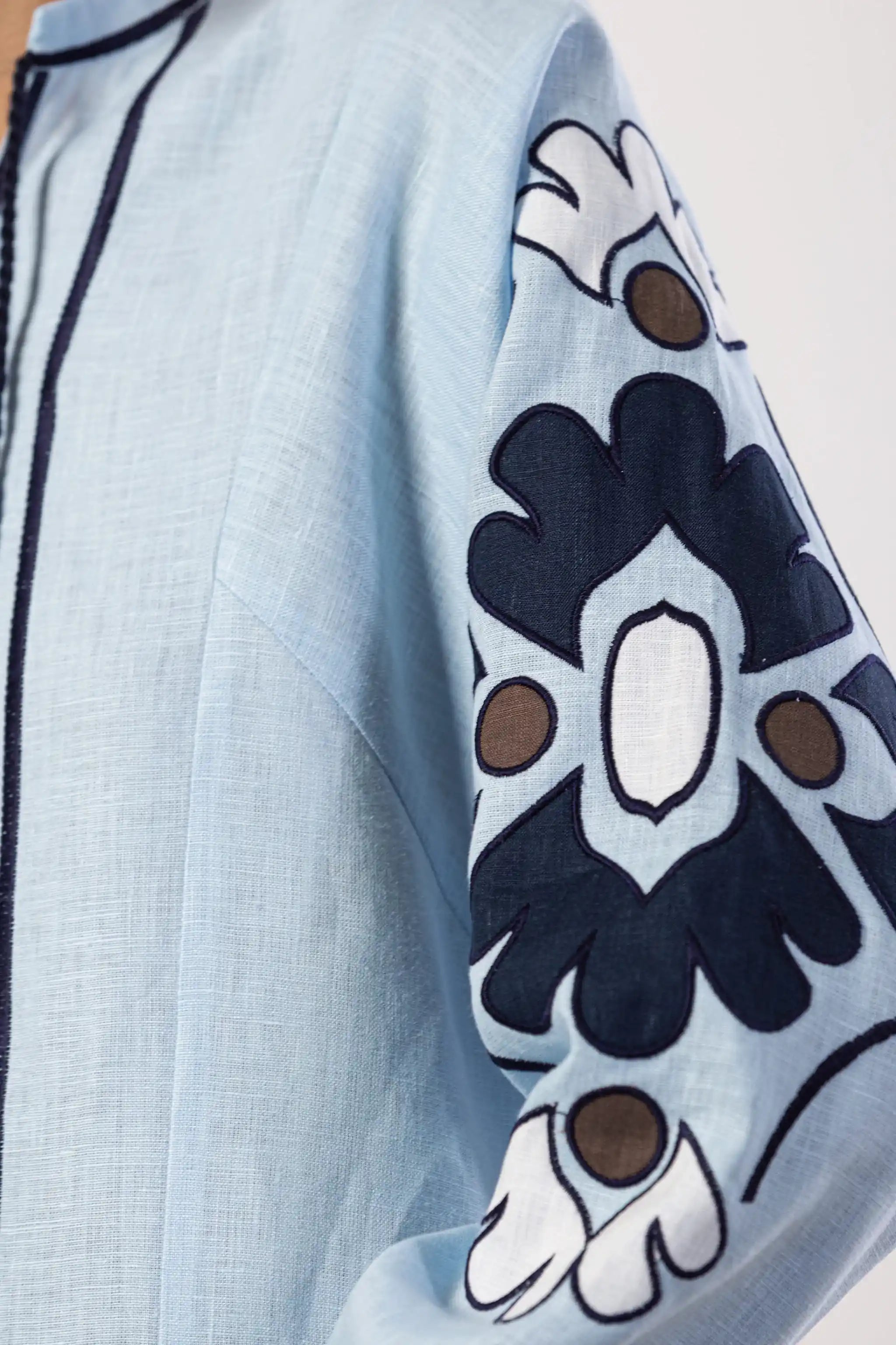 Close-up of a light blue linen garment sleeve with dark navy floral embroidery, white leaf details, and small brown accents.