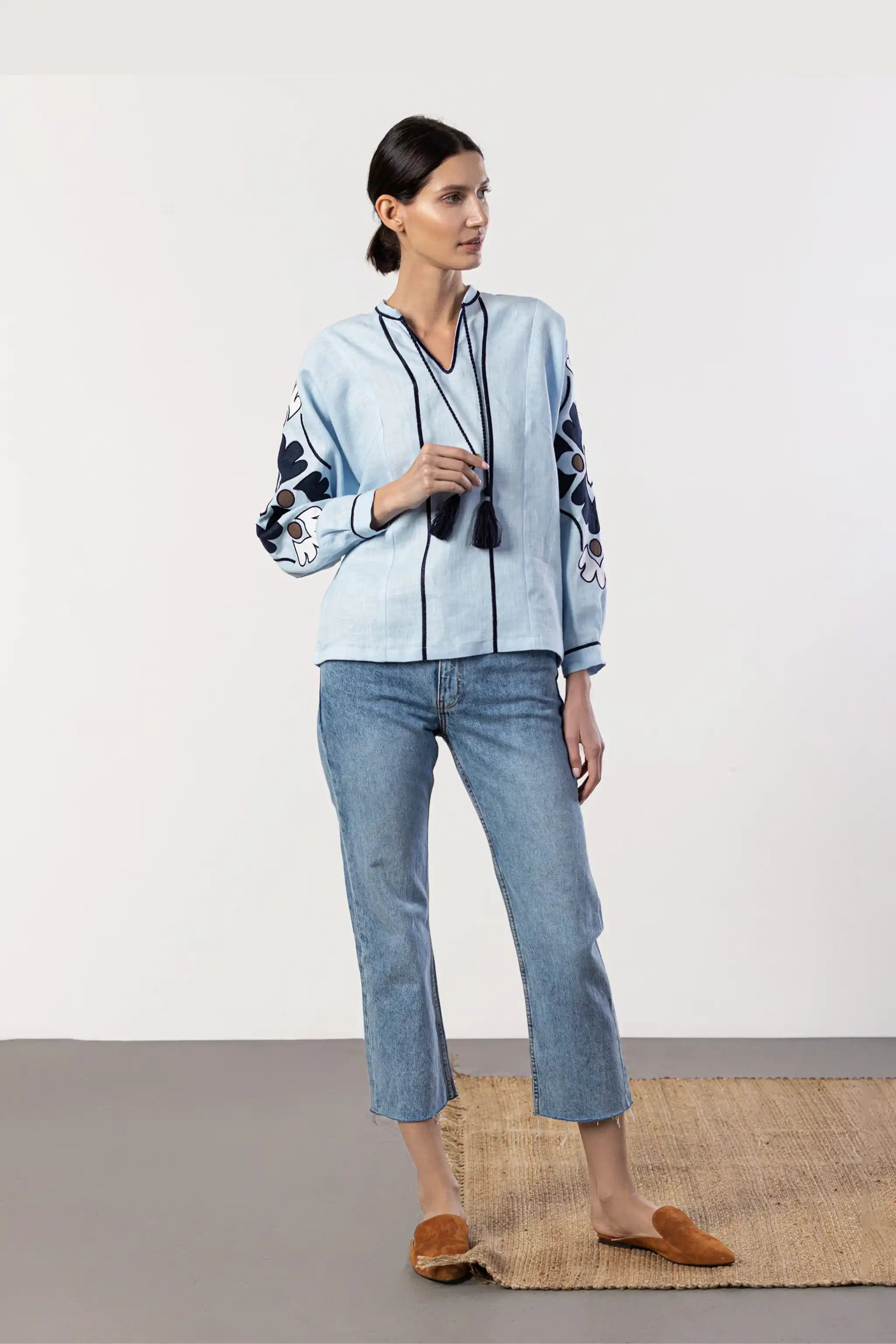 A woman models a light blue embroidered jacket with black trim, paired with blue jeans, brown mule slippers, and a black necklace, standing on a jute rug against a white studio background.
