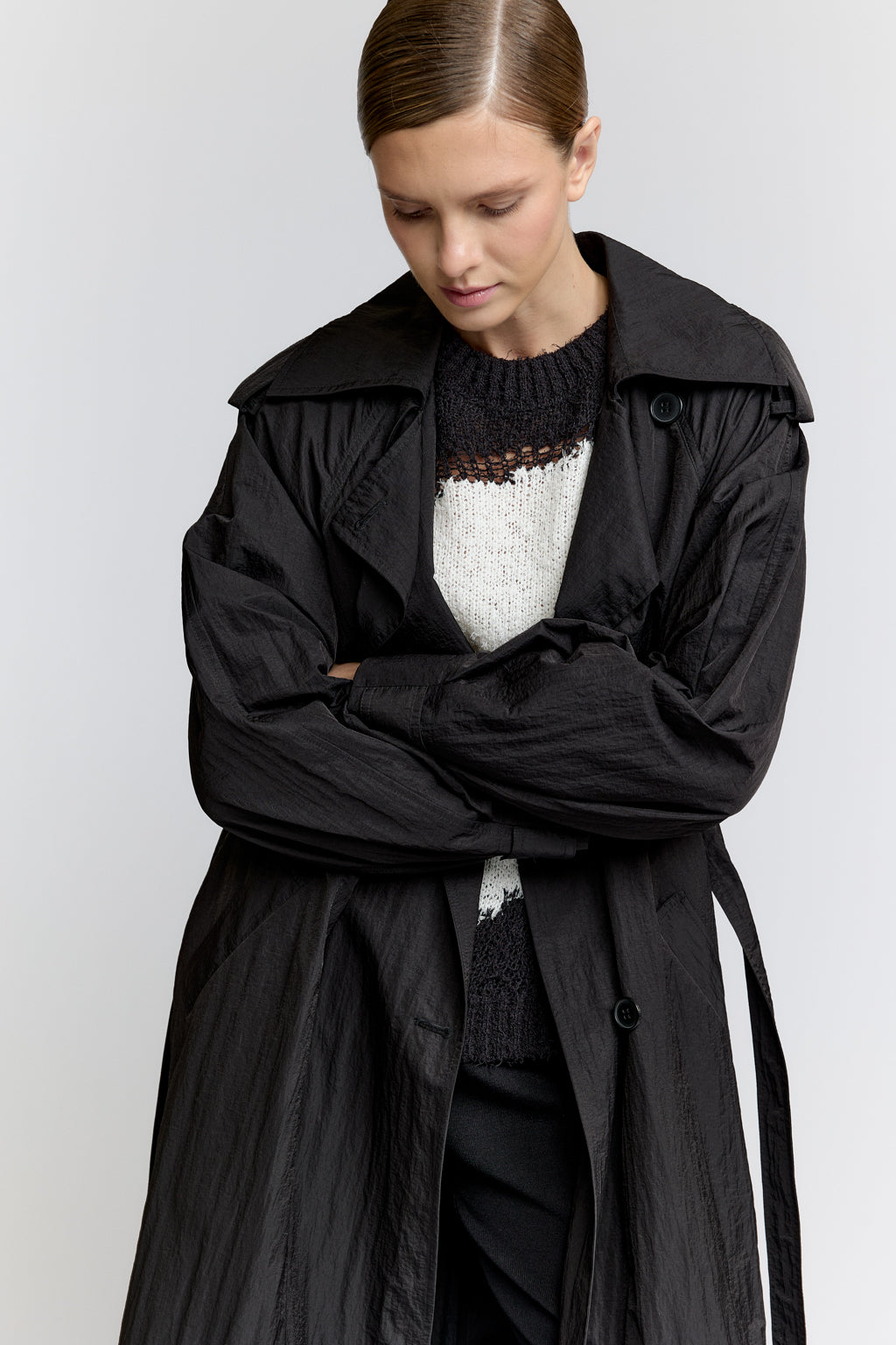Person wearing a black trench coat over a white sweater on a light gray background
