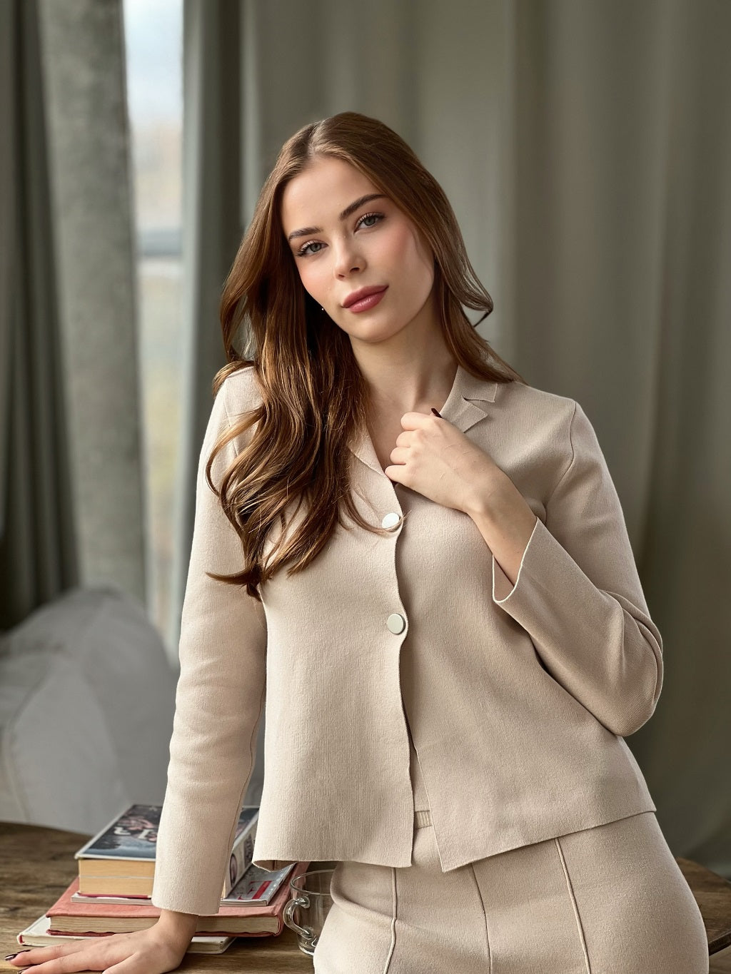 Woman wearing a beige suit sitting in a room with books and a window in the background