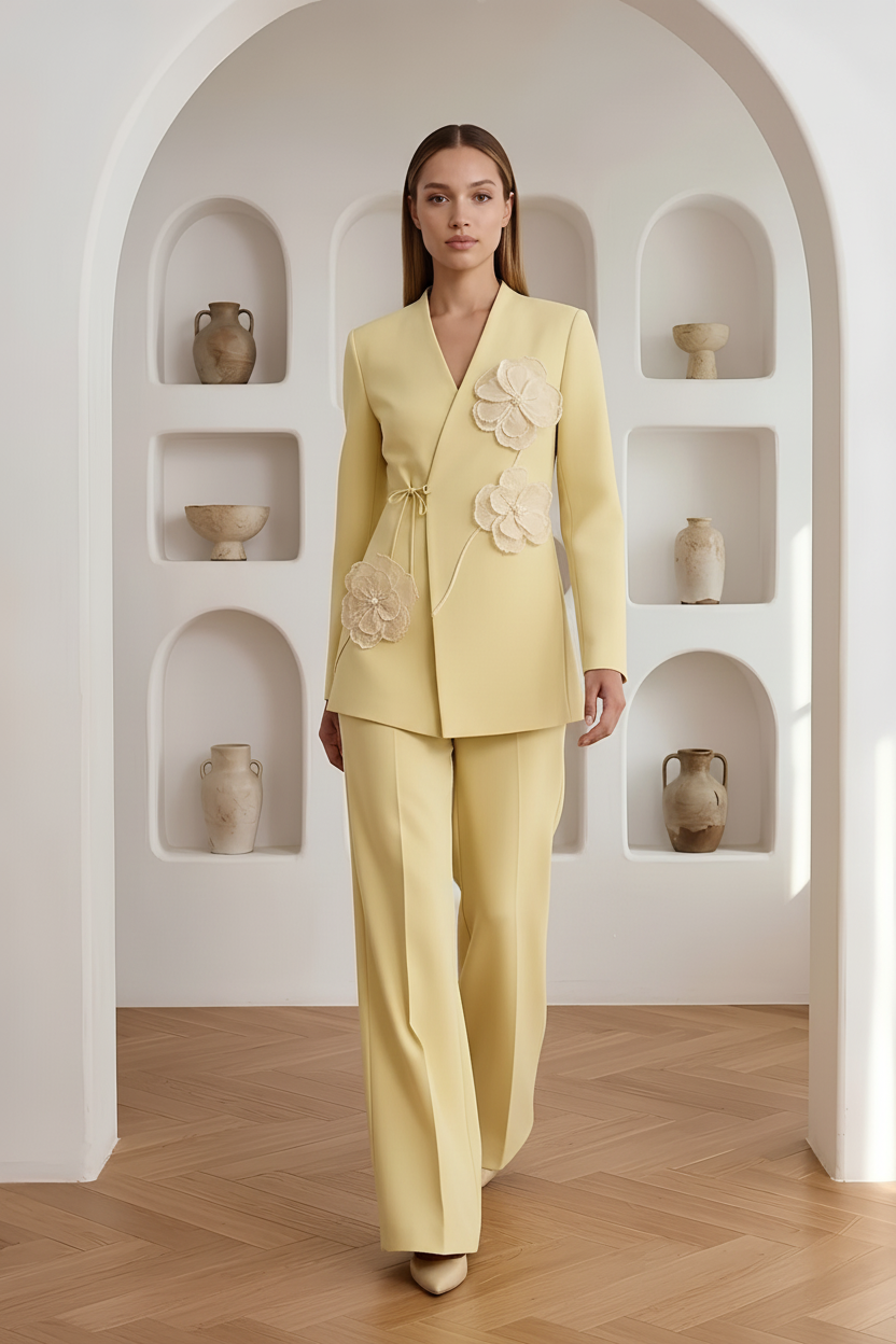 A model stands in a bright, minimalist room with arched alcoves and ceramic vases, wearing a pale yellow tailored suit with large floral appliqué details on the jacket and matching wide-leg trousers.