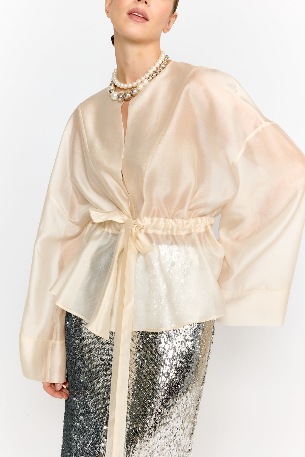 Woman wearing a beige sheer top with a pearl necklace and silver sequin pants on a white background