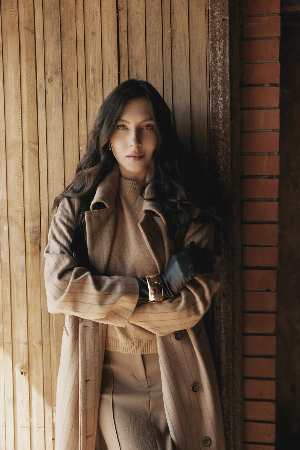 Woman in a beige coat standing against a wooden wall.