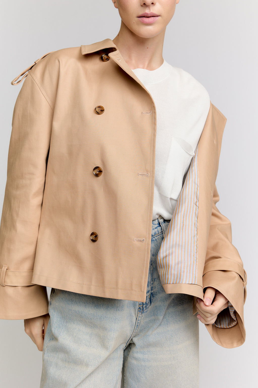 Person wearing a beige jacket over a white shirt with light blue jeans on a plain background