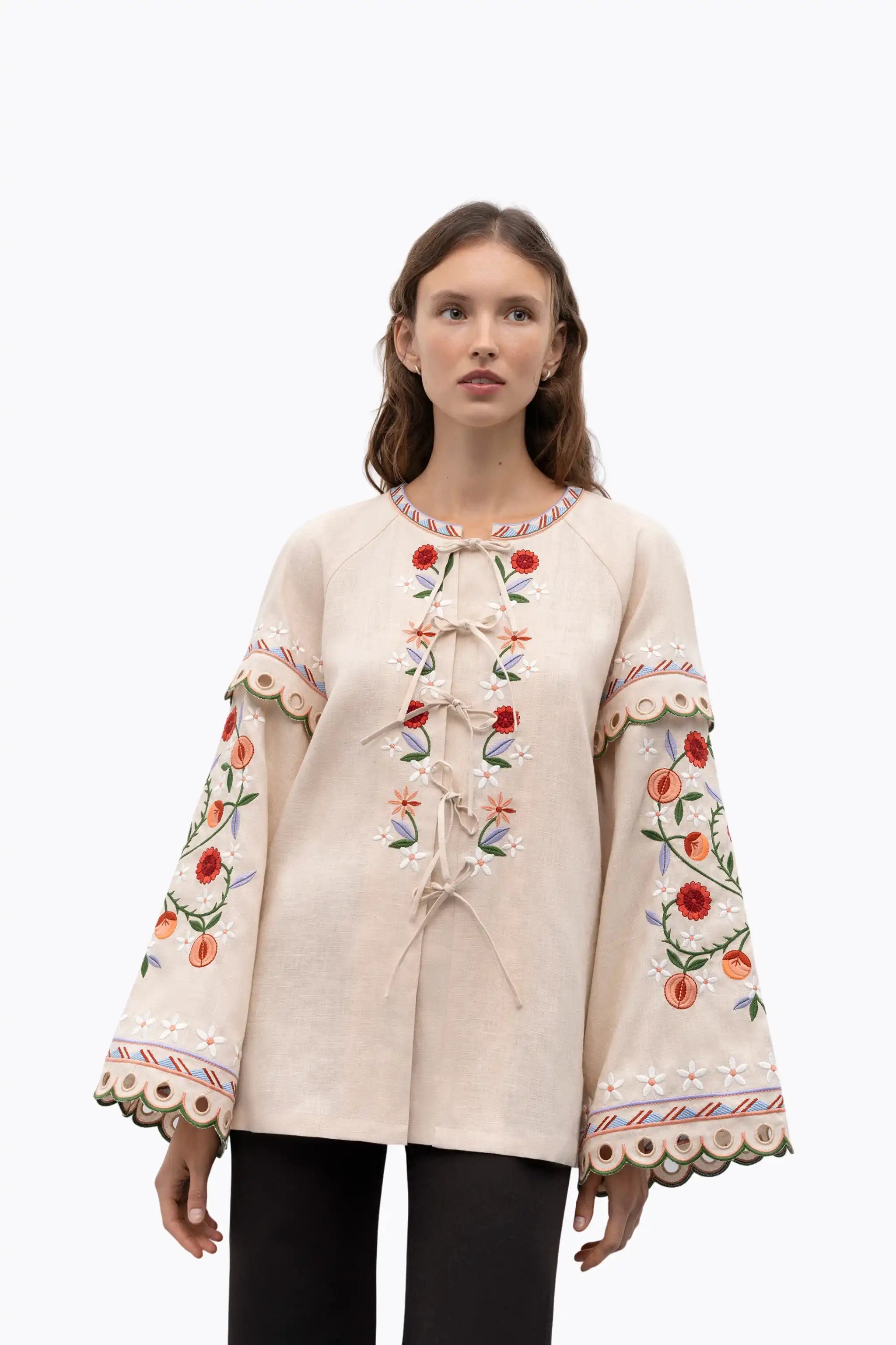 A model wearing a cream-colored linen jacket with colorful floral embroidery on the front and sleeves, paired with black pants, shot against a solid white background.