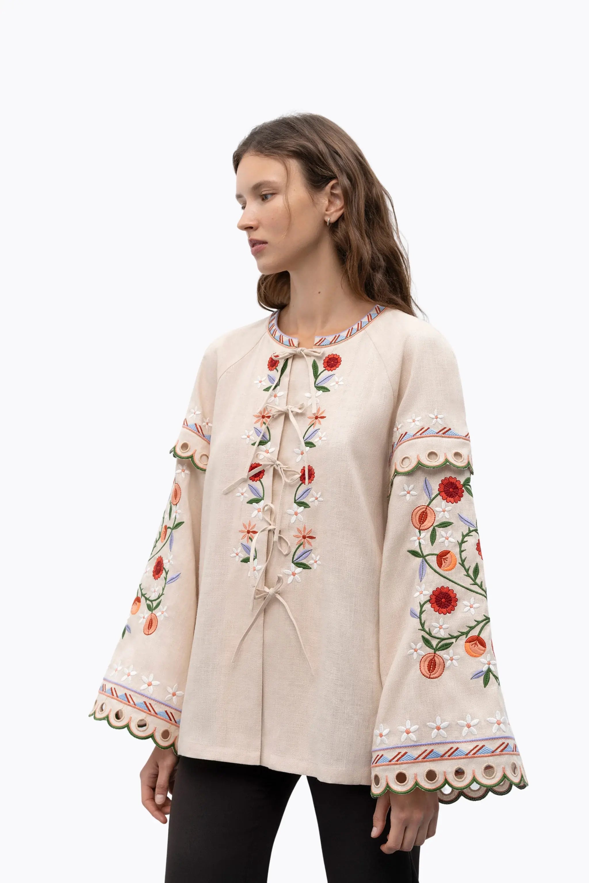 A woman wearing a cream-colored embroidered jacket with long bell sleeves, floral patterns, and front tie closures, photographed against a plain white studio background.
