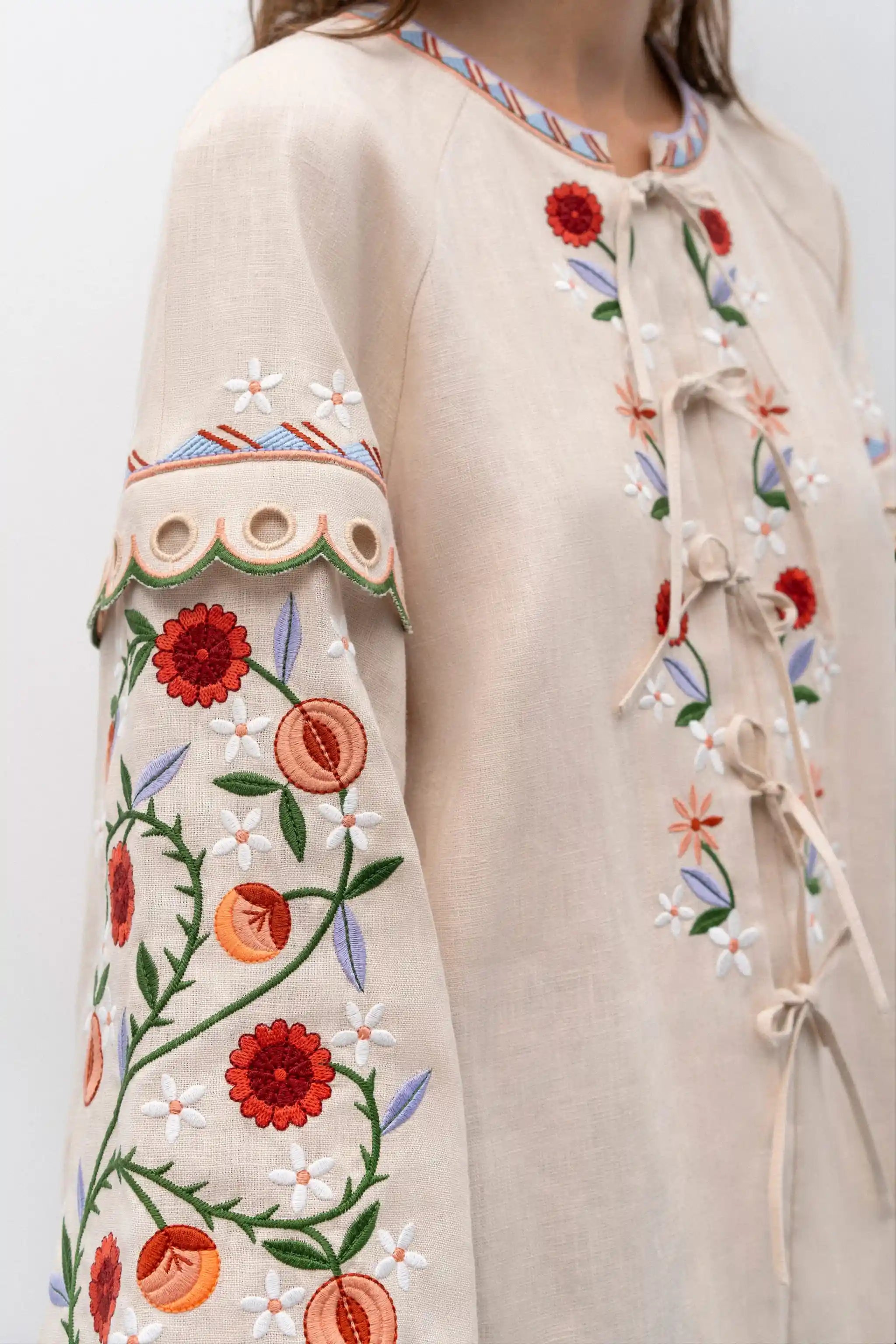 Close-up of a beige linen tunic top with colorful floral embroidery on the sleeve and front, featuring tie closures and eyelet detailing.