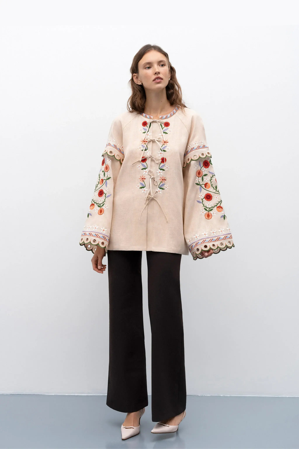 A woman modeling a beige long-sleeve blouse with colorful floral embroidery down the front and on the sleeves, paired with black wide-leg pants and beige pointed heels, set against a plain light gray studio background.