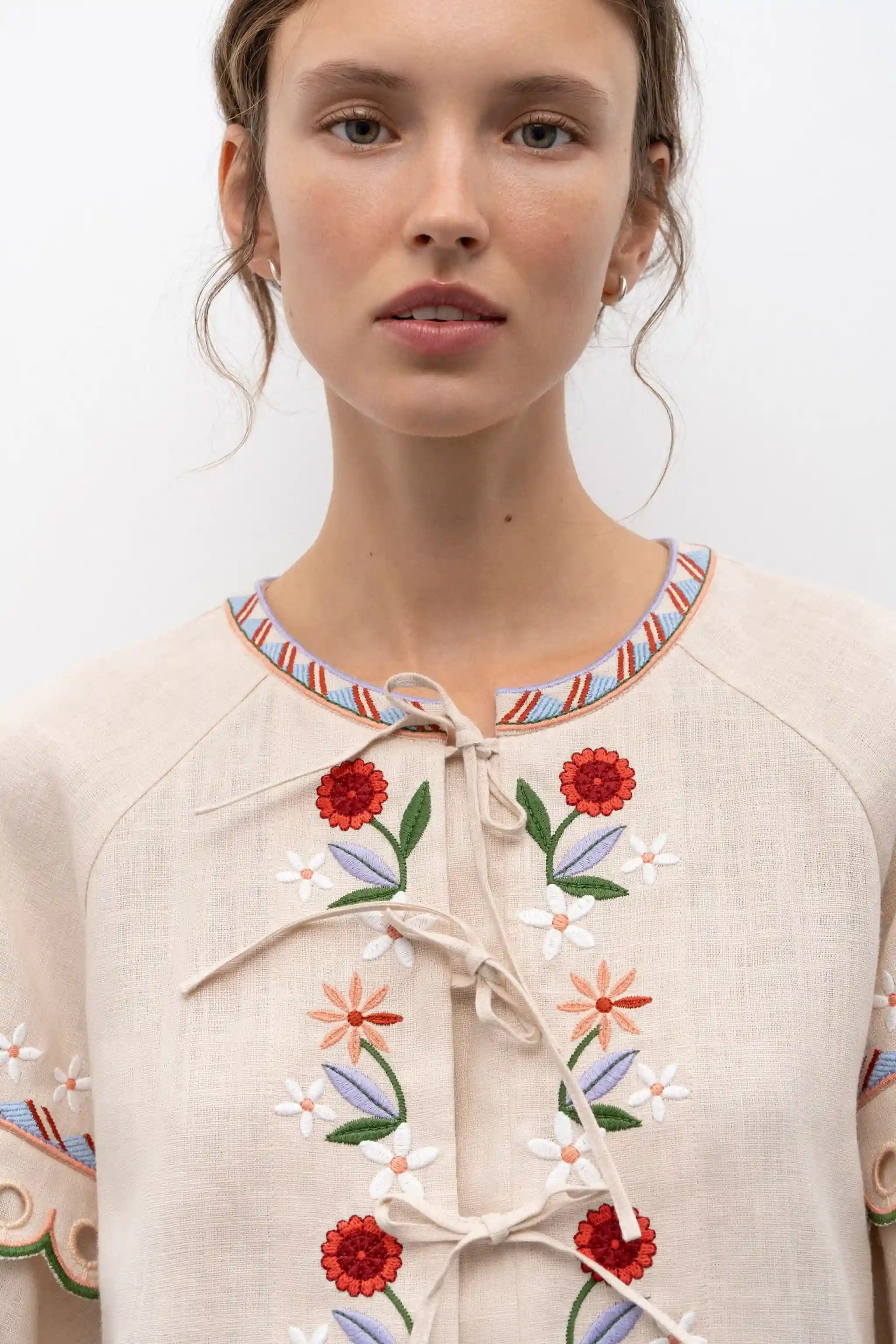 A close-up of a model wearing a light beige linen top with colorful floral embroidery and tie-front closures.