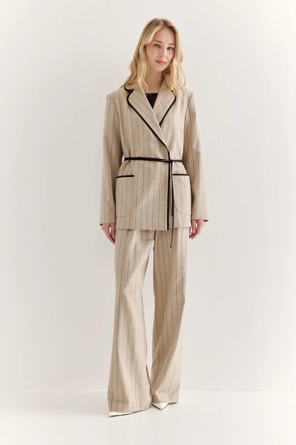 A woman modeling a beige pinstripe two-piece suit with black trim, featuring a wrap-style blazer with a tie belt and matching wide-leg trousers, set against a plain white studio background.