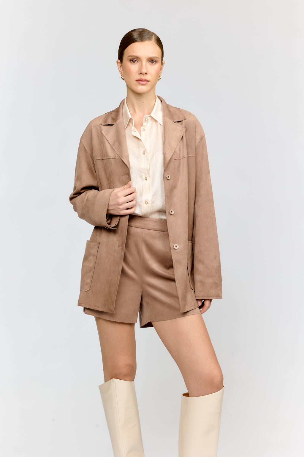 Woman wearing a beige jacket, shorts, and white boots on a light gray background