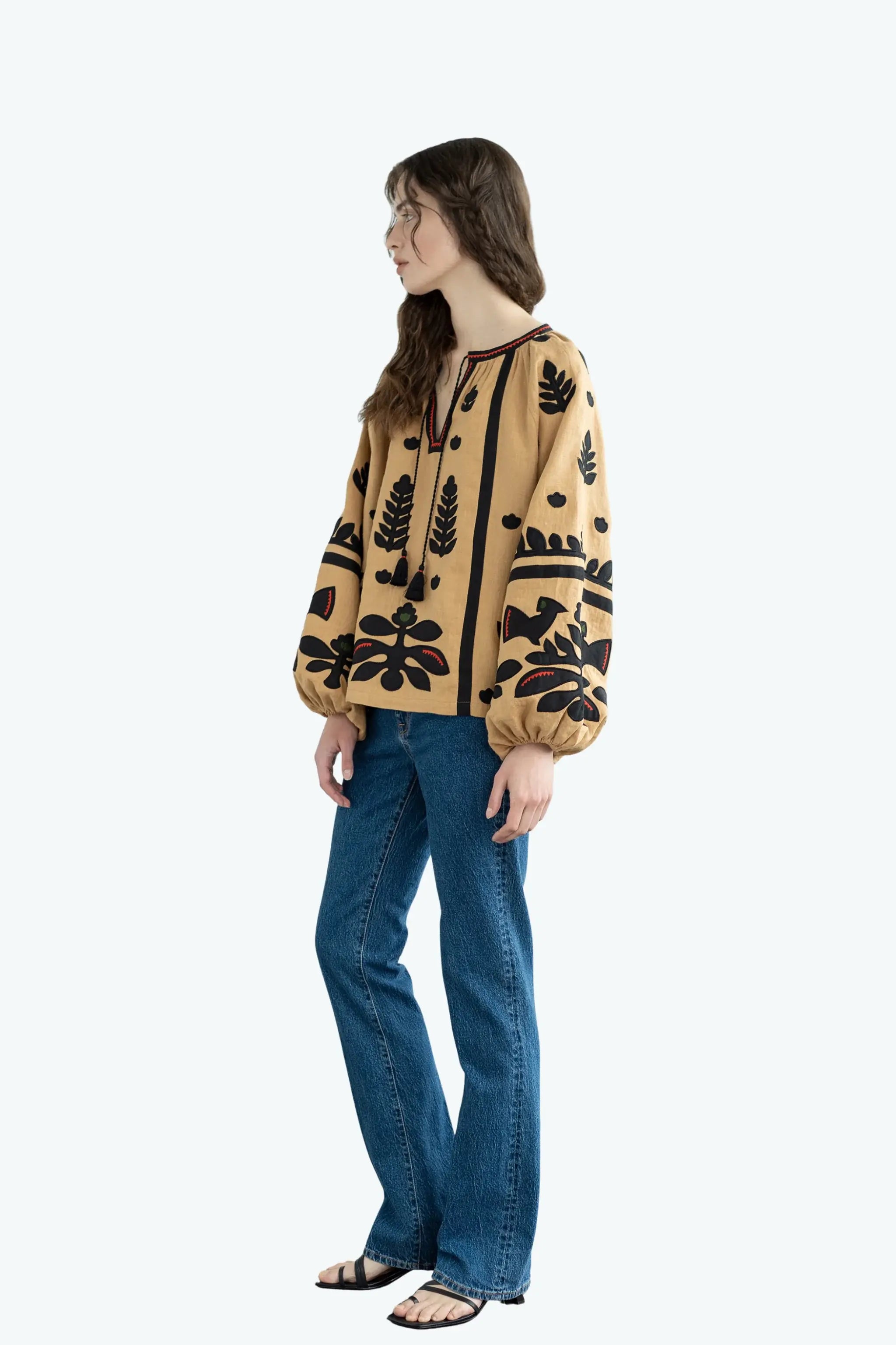 A woman modeling a beige long-sleeve blouse with black botanical and abstract patterns, paired with blue jeans and black sandals, shown from a side profile against a light gray background.