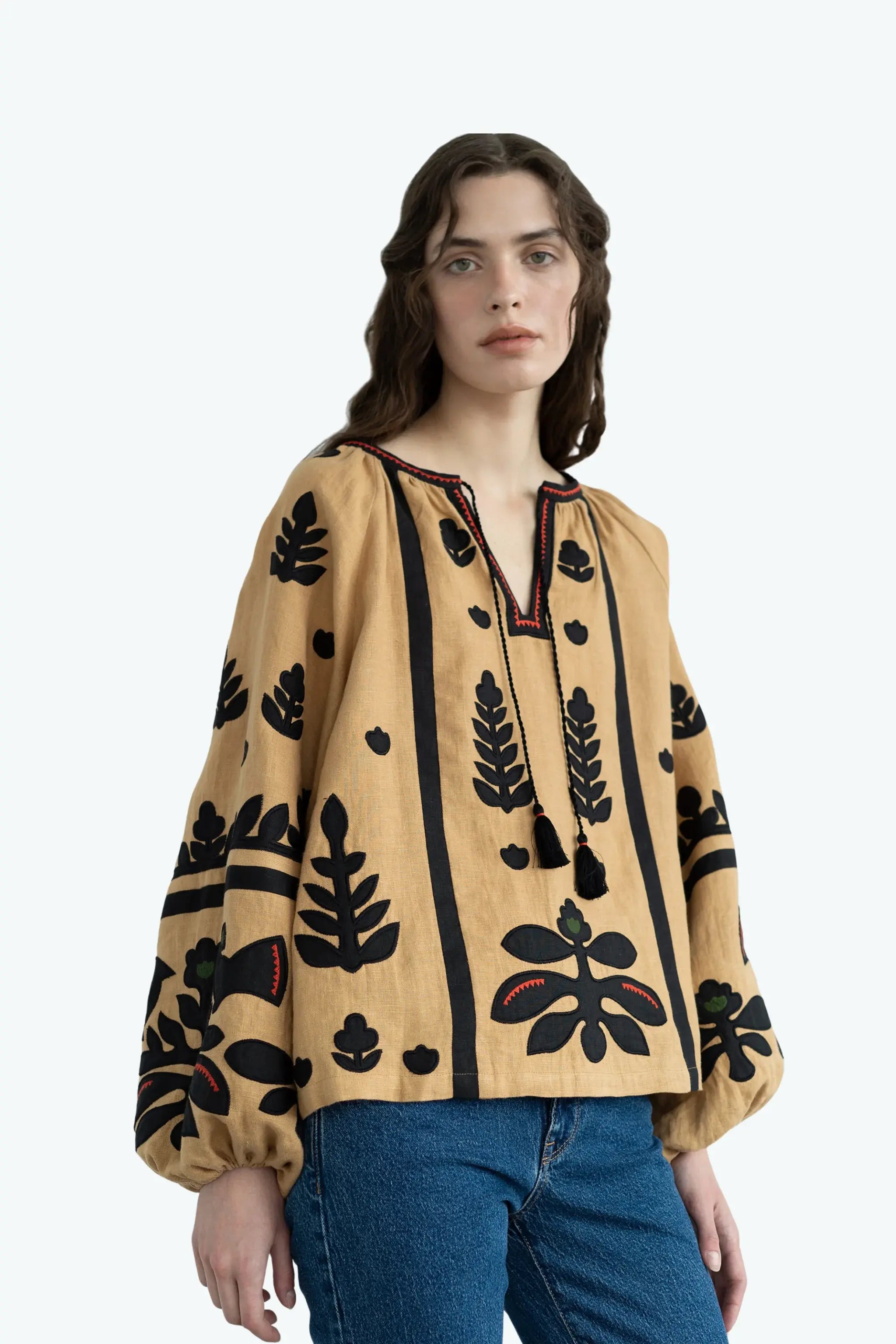 A woman wearing a beige long-sleeve blouse with black embroidered botanical patterns and blue jeans, photographed against a plain light gray studio background.