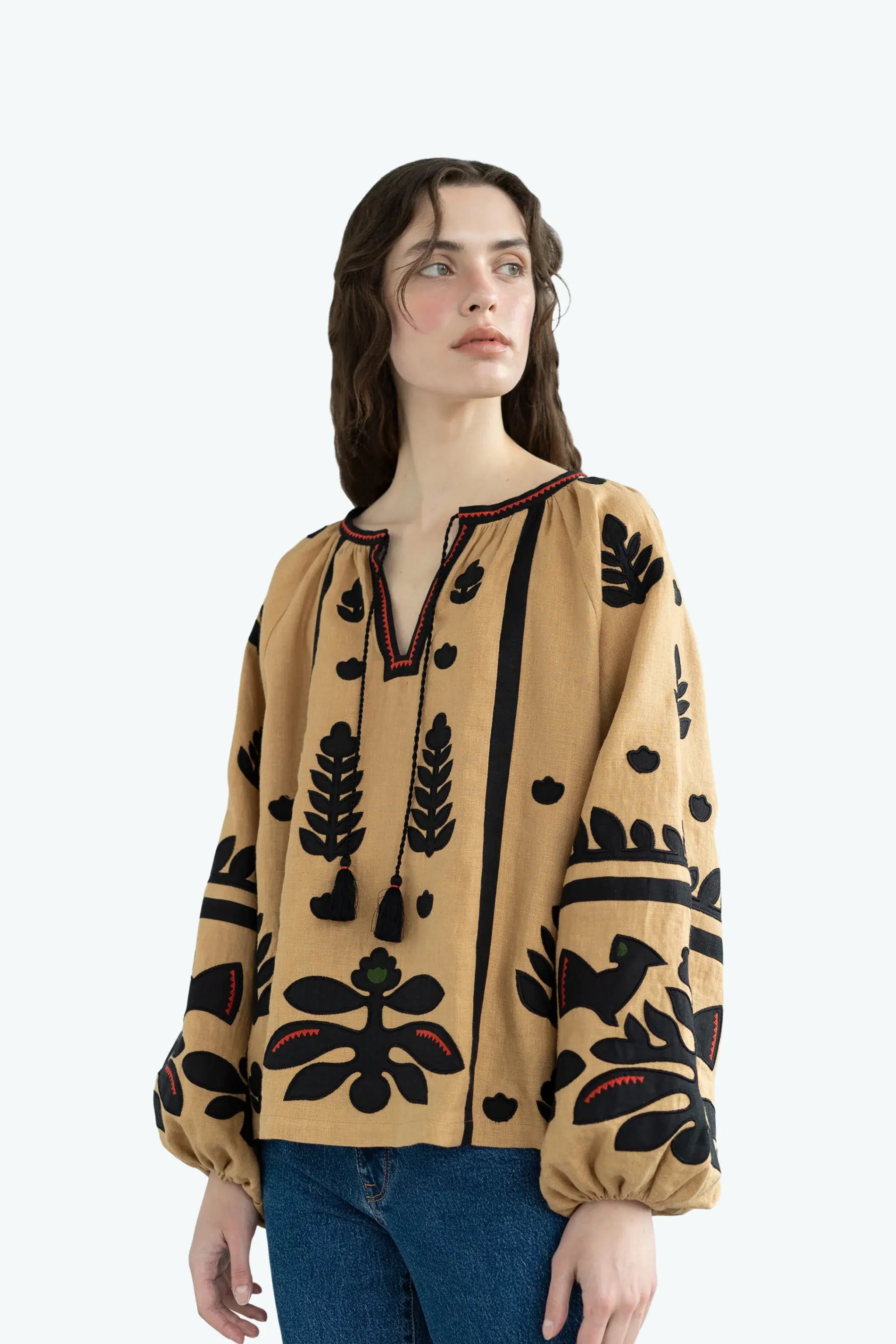 A woman modeling a beige long-sleeve blouse with black embroidered botanical and geometric patterns, paired with blue jeans, against a light gray studio background.